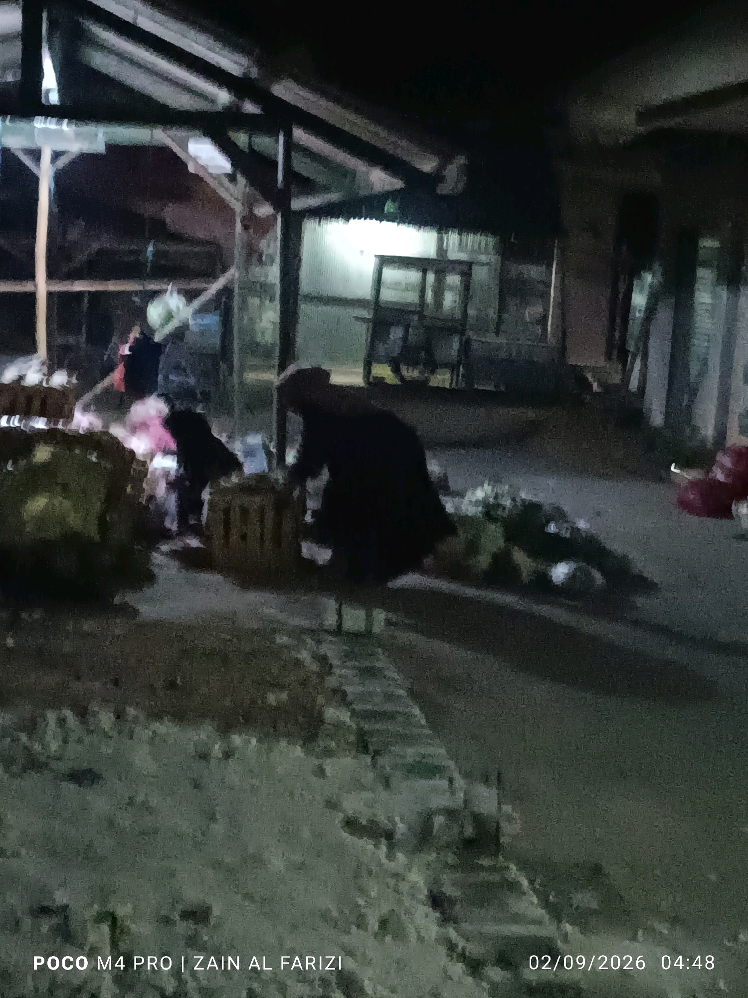 The image depicts a nighttime scene possibly in Arosbaya, Indonesia, focusing on an outdoor area with a rustic, open-sided structure. The lighting suggests artificial illumination, casting shadows and creating a somewhat grainy effect. In the foreground, what appear to be large, roughly shaped objects, possibly natural materials like wood or produce, are visible. To the left of these, a dark figure, likely a person, is partially obscured and appears to be engaged in some activity. Further into the scene, more wooden crates and possibly bundles of produce or materials are scattered. In the background, the structure shows a corrugated metal roof supported by wooden beams, and a glimpse of what might be shelves or stalls. The overall impression is of a market or storage area at night.