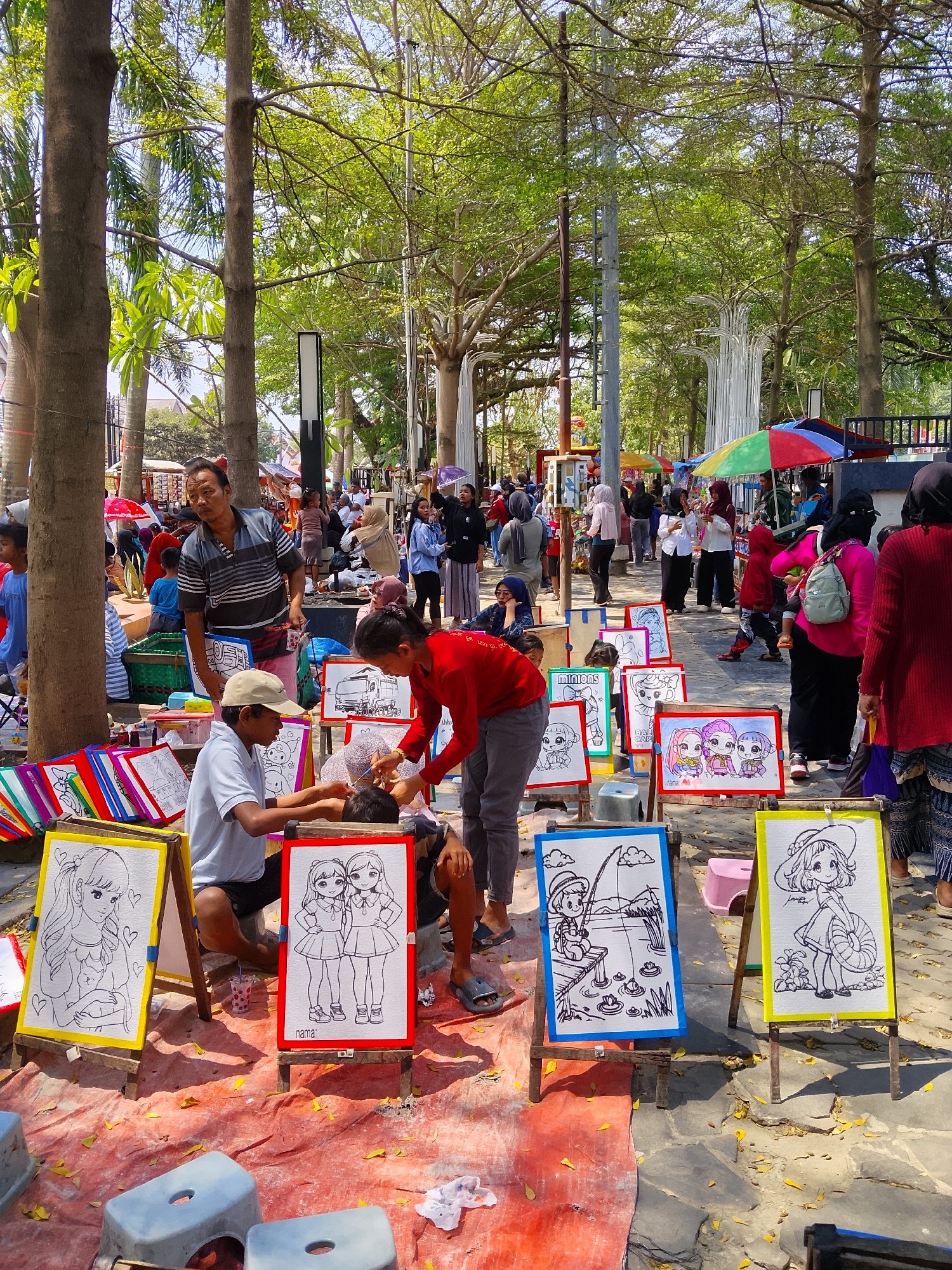 The image depicts an outdoor scene in Balun, Indonesia, likely a park or public space, on a sunny day. Several people are present, engaged in various activities. In the foreground, there are easels displaying black and white drawings of cartoon characters, some of which appear to be portraits or coloring pages. Two individuals, a man and a boy, are actively drawing or coloring on these displays. The man, wearing a cap and t-shirt, is focused on his work, while the boy, in a red shirt, seems to be assisting or interacting with him.
Behind them, more people are milling about, some observing the artists, while others are walking or socializing. Colorful umbrellas are visible in the background, suggesting a market or festival atmosphere. The environment is characterized by large trees providing shade, with dappled sunlight filtering through the leaves. The ground is covered with a reddish tarp and scattered yellow leaves, indicating a possibly dusty or natural setting. Overall, the scene captures a moment of artistic activity and community gathering in a public outdoor space.