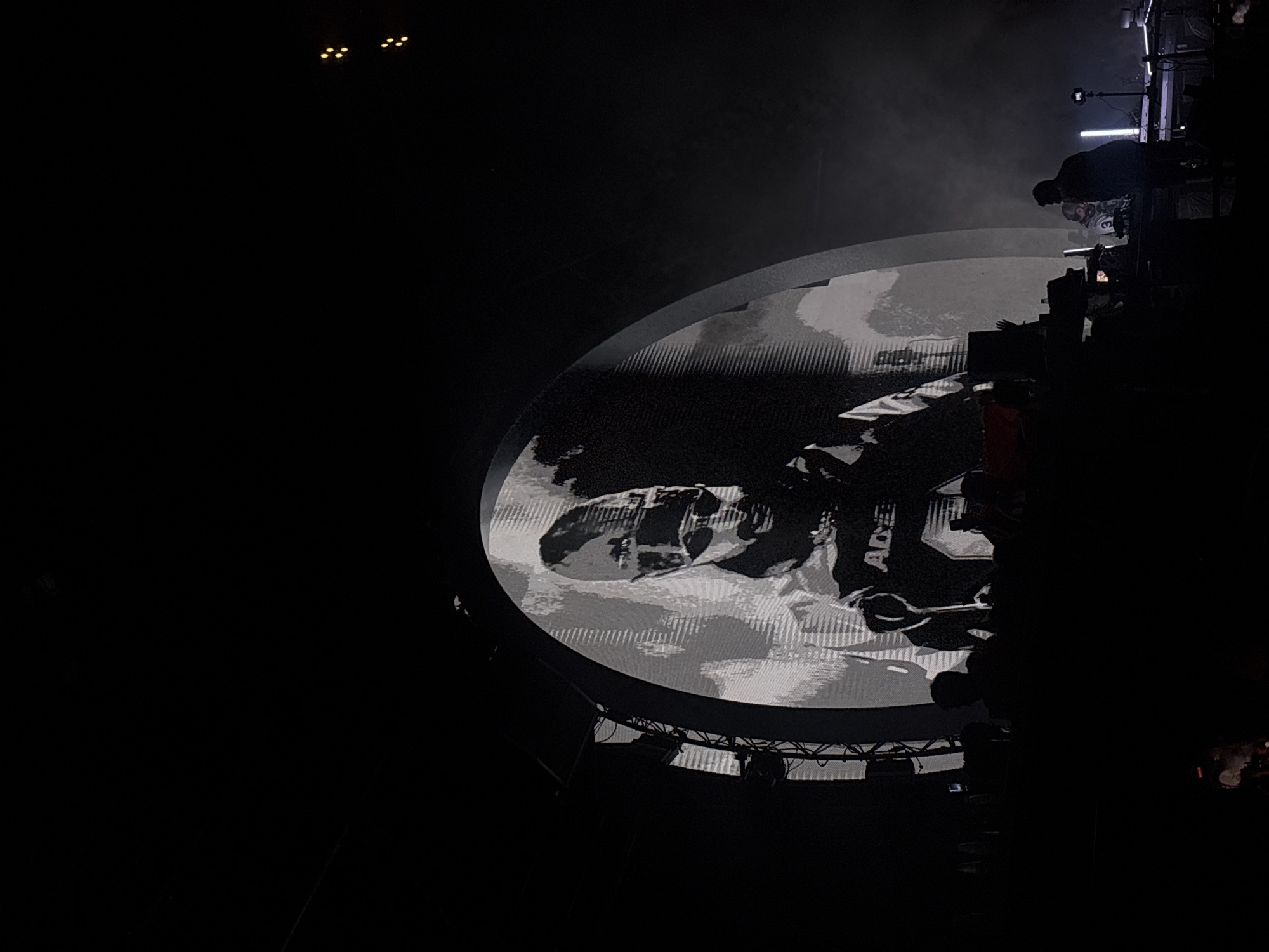 An indoor concert stage is set up in Sofia, Bulgaria, with a large, oval screen displaying a black and white image of a person wearing a hat and sunglasses, possibly a performer or a DJ. The screen is illuminated, creating a bright focal point in the dimly lit venue. Below the screen, several figures are visible in silhouette, suggesting an audience or stage crew. The overall atmosphere appears to be dark, with some artificial lighting contributing to the mood. The presence of what appears to be a DJ setup and stage lighting indicates a performance is underway or about to begin.