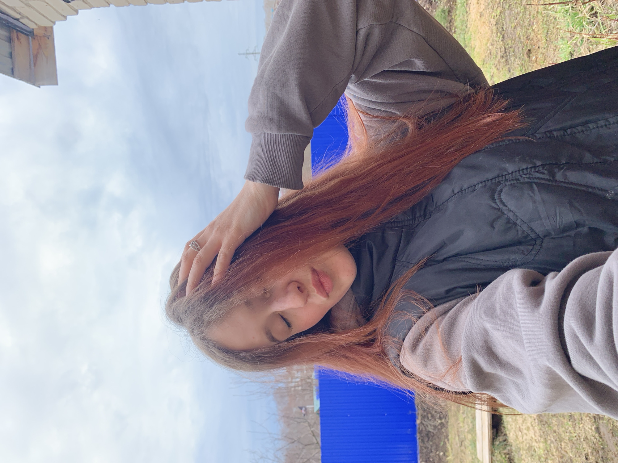 A woman with long red hair is depicted outdoors, likely in a backyard setting, suggested by the presence of grass and a fence, potentially located in Фоки, Russia. She is wearing a gray sweatshirt and a dark vest. Her eyes are closed, and one hand is resting on her head. The sky is visible in the background. A building is partially visible in the upper left corner.
