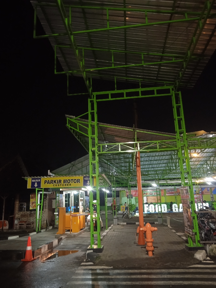The image depicts a nighttime scene at the entrance of a facility, identified as a "FOOD GARDEN" in Kecamatan Kartasura, Indonesia. The most prominent feature is a large, brightly painted green metal structure that forms an overhead canopy and an entrance archway, supported by an intricate framework of beams. The canopy has a corrugated metal roof.
On the left side of the entrance, a well-lit parking booth with yellow and white accents is visible. A sign above the booth clearly states "PARKIR MOTOR CUSTOMER" (Motorcycle Parking Customer), accompanied by a blue arrow pointing downwards to indicate the entrance. Another sign, partially visible, likely signifies the exit. An orange and white barrier arm is part of the parking system. An orange traffic cone rests on the ground nearby, which shows puddles, suggesting recent rain.
A person, possibly a parking attendant, is visible near the booth in a light-colored uniform. Further inside the facility, additional people and several motorcycles can be vaguely seen.
In the center of the main entrance, a distinct bright orange, pipe-like structure, resembling either a decorative bollard or a fire hydrant, stands out. Beyond the immediate entrance, more green metal canopies and structures extend into the dimly lit background, leading towards a larger area where a prominent sign displays "FOOD GARDEN." Artificial lighting illuminates the structures and the immediate ground, contrasting with the dark night sky. The scene captures the functional operation of a commercial establishment's motorcycle parking entrance after dark.