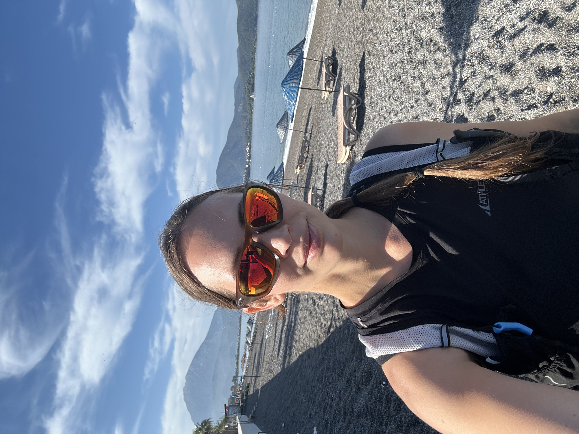 The image is a selfie of a woman on a pebble beach in Fethiye, Turkey. She is wearing sunglasses with orange lenses and a black athletic shirt with white mesh panels. Her brown hair is pulled back. The beach is gray and rocky, with blue and white striped umbrellas and lounge chairs in the background. In the distance, there is a mountain range. The sky is blue with scattered white clouds. Some buildings are visible along the coast behind the beach.