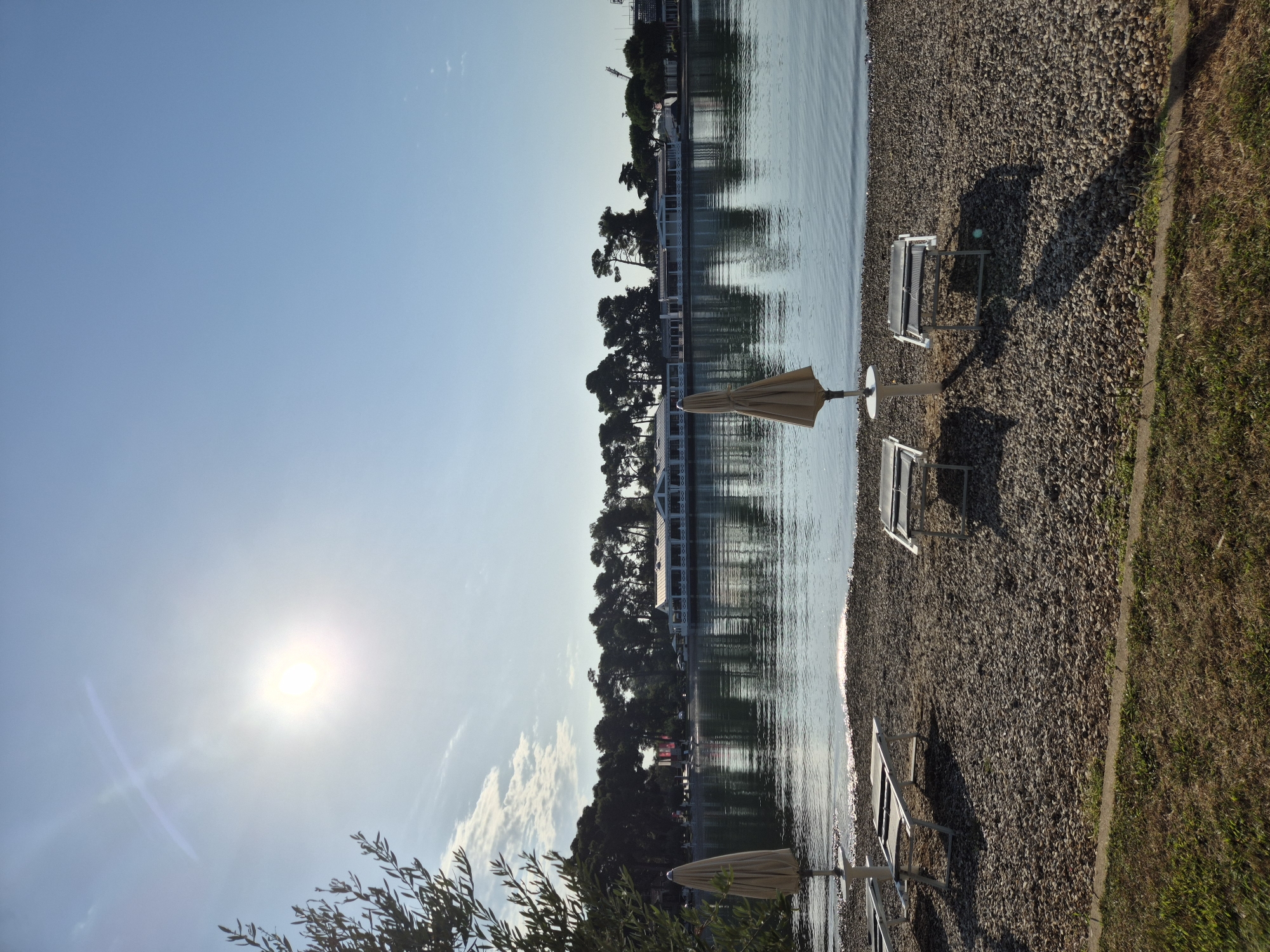 The image depicts a serene beach scene in Umag, Croatia, with the sun shining brightly in a clear blue sky. In the foreground, a pebbled beach is lined with several empty lounge chairs and closed umbrellas, suggesting a quiet, pre-season or post-season atmosphere. The calm, reflective water of the Adriatic Sea gently laps at the shore. Across the water, a long, white building with numerous windows and a covered porch is visible, backed by a dense line of dark green trees. This establishment appears to be a restaurant or hotel, characteristic of the coastal resorts found in Umag. The overall impression is one of tranquility and natural beauty, with the architecture of the building subtly complementing the natural landscape.