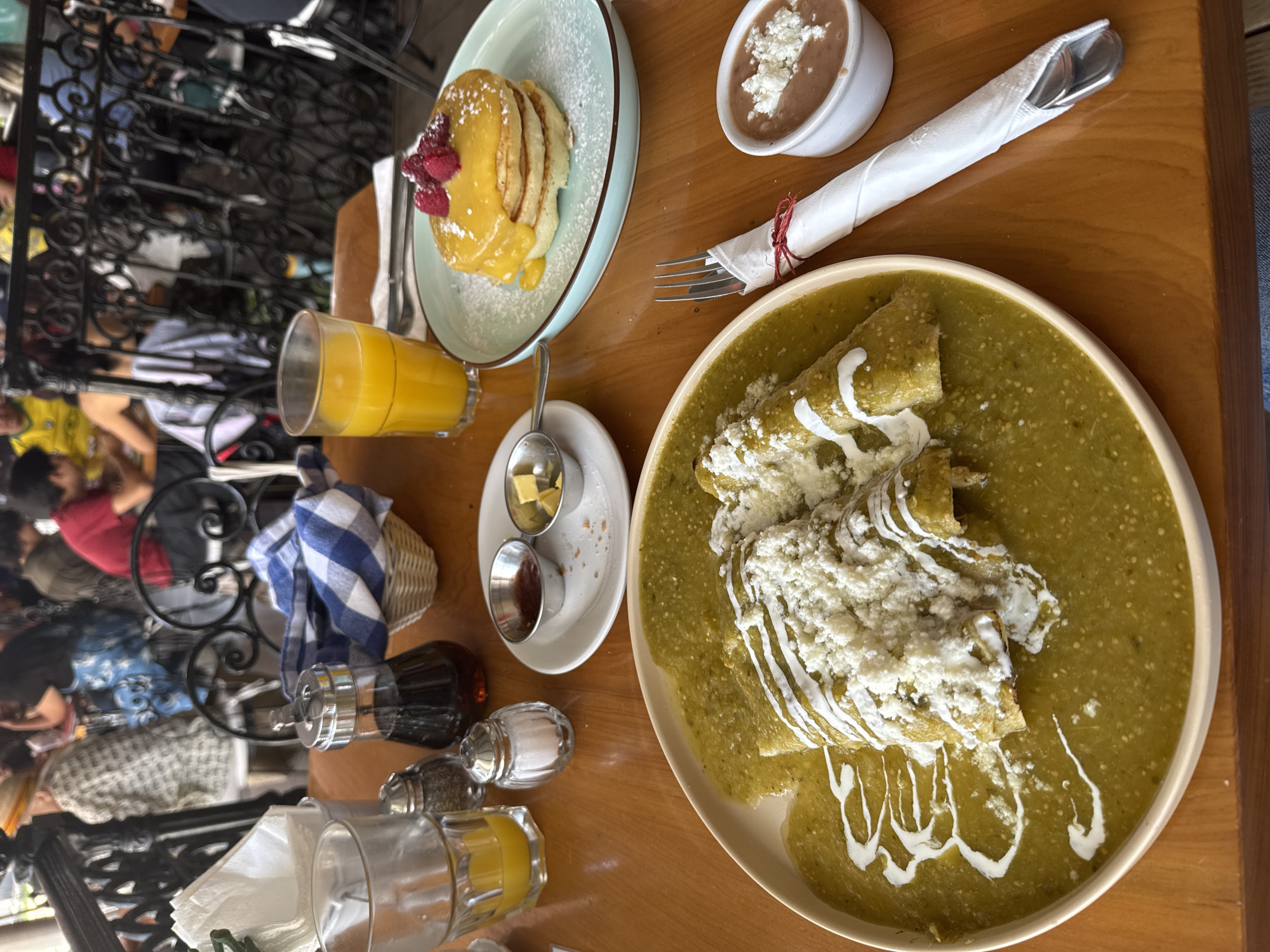 The image shows a table at a restaurant in San Miguel de Allende, Mexico, set with a breakfast spread. The table is wooden and features various items: a plate of enchiladas verdes topped with cream and cheese, a plate of pancakes with raspberries, a side of refried beans topped with cheese, a glass of orange juice, a small dish with butter and jam, and condiments like syrup, salt, and pepper. Cutlery wrapped in a napkin is also present. The setting appears to be outdoors, as parts of people and a wrought iron barrier can be seen in the background. People are seated at a nearby table.