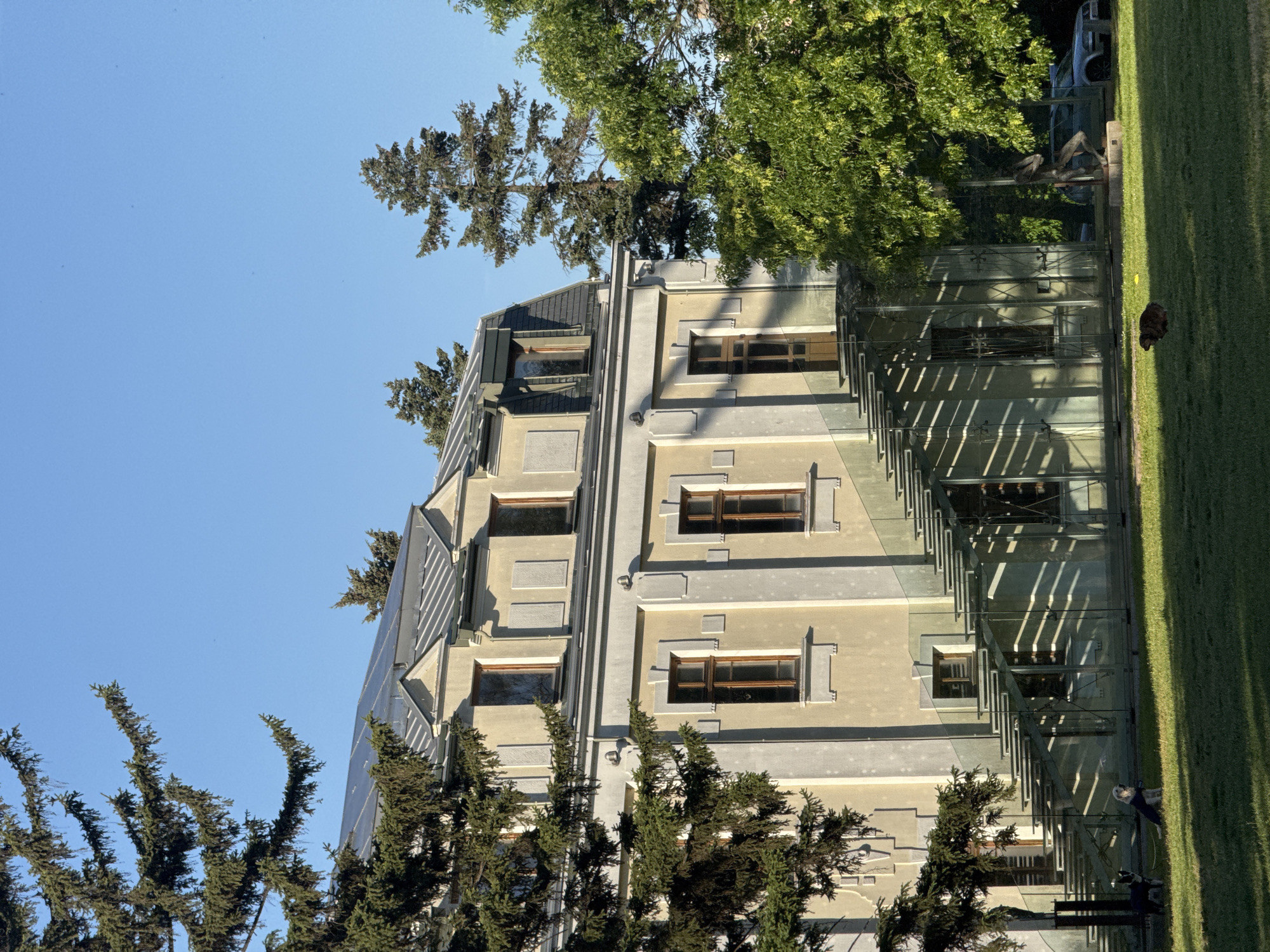 An exterior shot of a modern building with a beige facade and gray accents in Sofia, Bulgaria, under a blue sky. The building has multiple rectangular windows with brown frames. Adjacent to the building is a glass structure with metal supports and long diagonal shadows stretching across the lawn. Evergreen trees are framing the building. On the lawn, there is a tree and sculpture. A car parked in the distance.