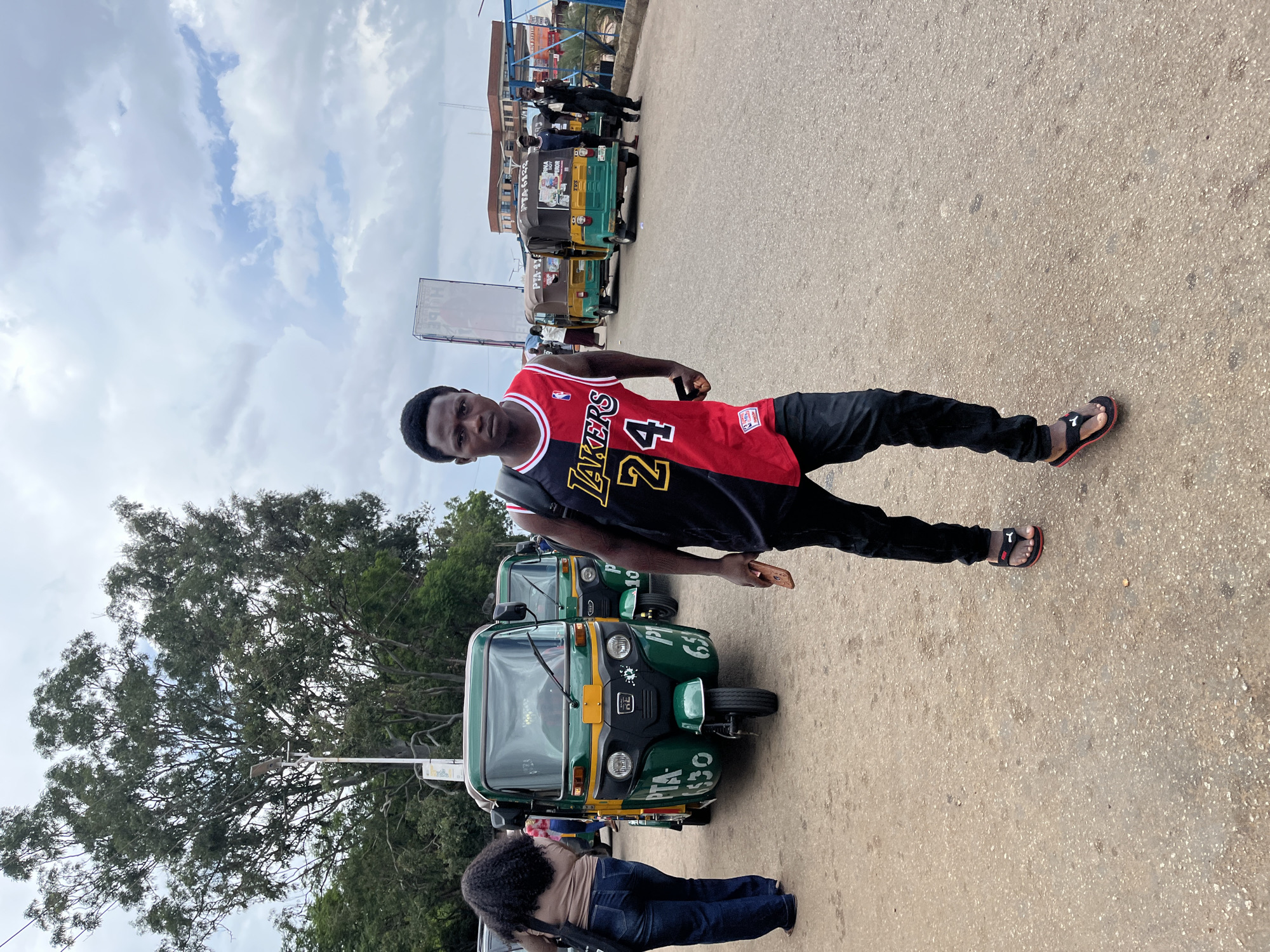 A young man wearing a Los Angeles Lakers basketball jersey with the number 24 and black pants walks towards the camera on a paved street in Jos, Nigeria. He is holding a mobile phone in his right hand. The street is lined with several green and yellow auto rickshaws, known locally as "Keke NAPEPs." In the background, a large tree with dense foliage is visible, along with a tall white billboard. The sky is overcast with scattered clouds. A woman with long, dark curly hair wearing a light brown top and blue jeans is walking away from the camera on the left side of the frame, near one of the auto rickshaws. Other people are present in the background, near the auto rickshaws and buildings. The overall scene depicts a busy urban environment, likely a transportation hub.