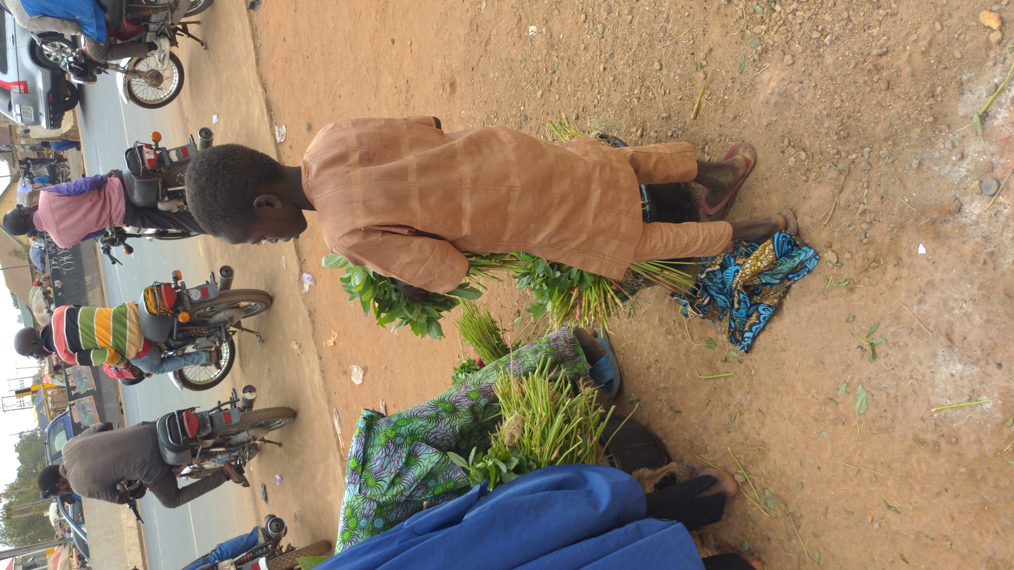 Ado, Nigeria: A young person in a tan outfit lies on the ground amidst bundles of green plants and stalks. Their head is turned towards a line of motorcycles parked along the side of a street. Other people are on the motorcycles, facing in the same direction. Several are wearing long-sleeved shirts, and one has a striped shirt. A blue and green patterned cloth lies near the person. The ground is dirt and small pebbles. Cars are also visible in the background.