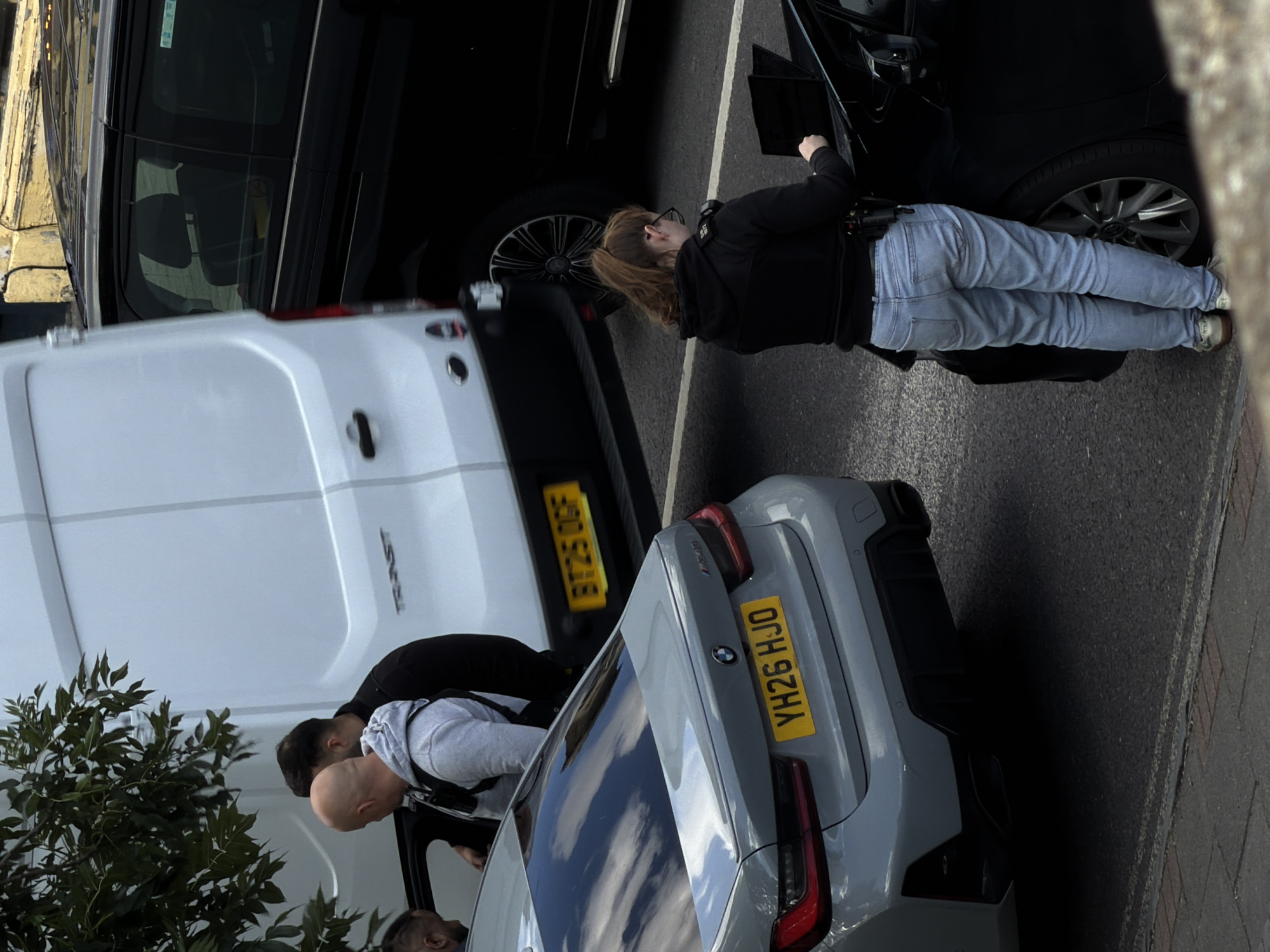 The image depicts an outdoor scene, likely on a street in London, United Kingdom, based on the presence of vehicles with UK license plates. In the foreground, a grey BMW sedan is parked. Behind it, a white transit van is visible, and to the right, a black van or SUV. A woman wearing light blue jeans, a black hoodie, and glasses is standing next to the black vehicle, interacting with a laptop placed on the vehicle's hood. She appears to be wearing a uniform, indicated by patches on her sleeve and a utility belt. To the left of the BMW, a man in a grey hoodie and another man in a black jacket are near the open door of a vehicle, possibly the grey BMW or another vehicle not fully visible. The ground is paved, with a curb and sidewalk visible on the lower edge of the frame. The overall atmosphere suggests an official interaction or investigation taking place.