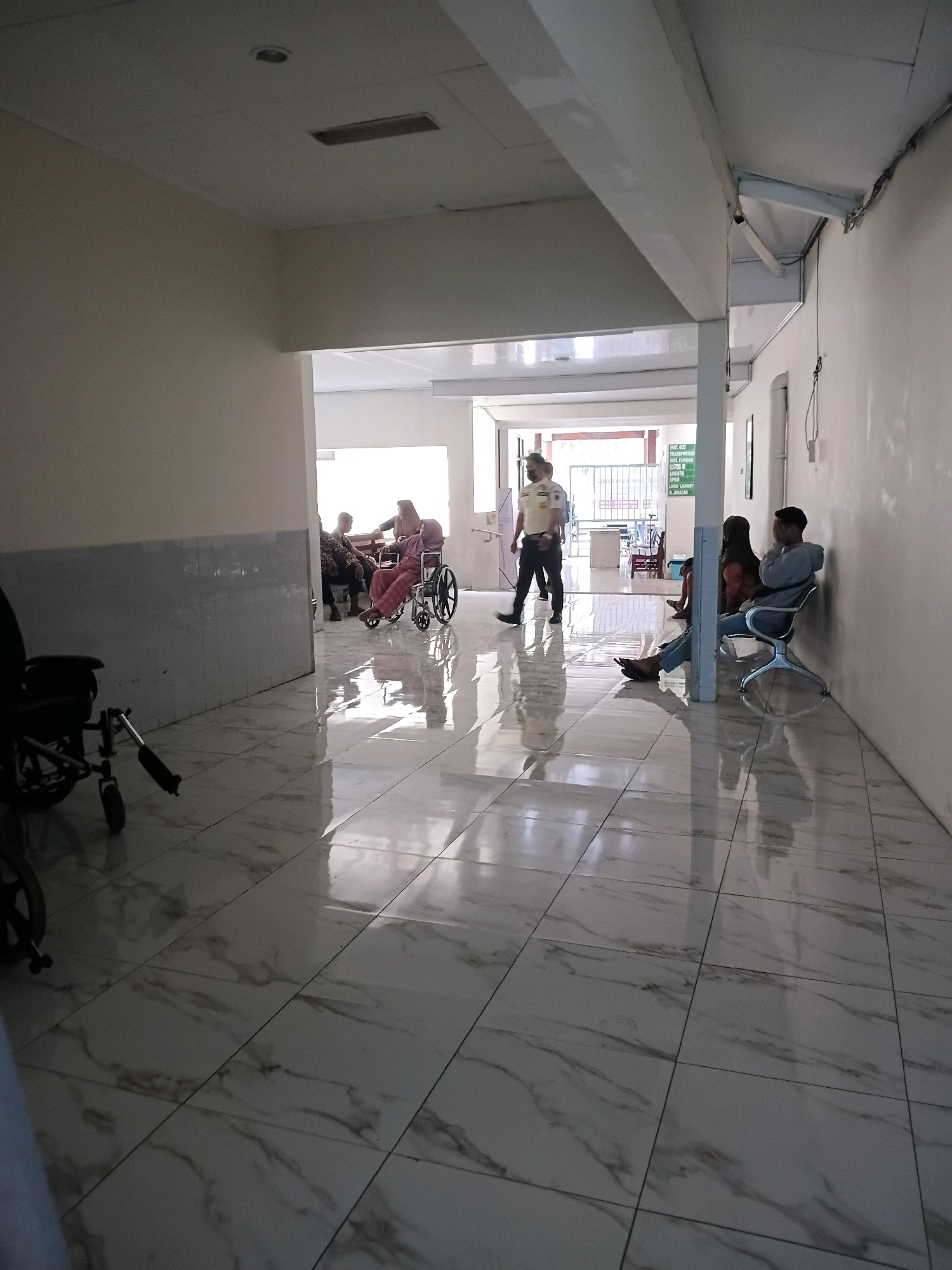 In a clean, modern hospital hallway with marble-tiled floors reflecting the bright overhead lighting, a security guard in a light-colored uniform walks towards the left, his face partially obscured by a mask. To his right, a man in a grey hoodie and jeans sits on a contemporary chair, his head bowed, while a woman in a hijab sits beside him. Further down the hallway, to the left, a woman in a pink headscarf and dress is seated in a wheelchair, conversing with another woman. In the background, more individuals are visible, some seated, suggesting a waiting area. A prominent sign with green lettering is mounted on a pillar. The hallway is illuminated by recessed ceiling lights and natural light streaming in from an unseen area at the end. The overall impression is of a tidy, functional hospital environment in Purwodadi, Indonesia, likely a waiting area or passage within the facility.
