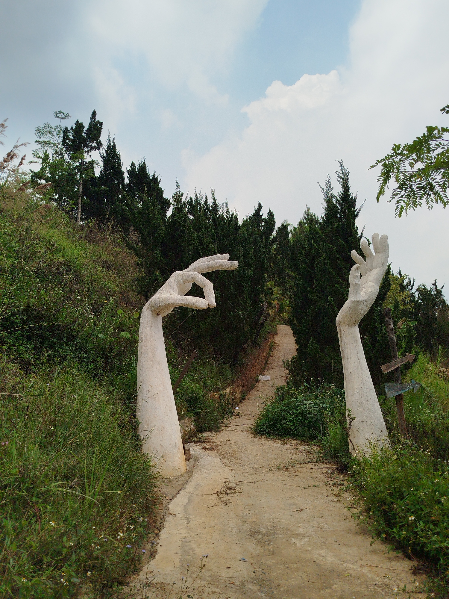 The image shows a pathway in what appears to be a garden or park in Da Lat, Vietnam. Two large, white concrete sculptures of hands act as an archway at the entrance to the path. The hand on the left side of the path has its index finger and thumb touching, creating a small opening. The other hand is open, facing upwards. The path is made of dirt or concrete and leads upwards into an area densely populated with evergreen trees. Lush green vegetation grows on either side of the path, obscuring the ground. A directional sign is visible on the right side. The sky is overcast with a mix of clouds.