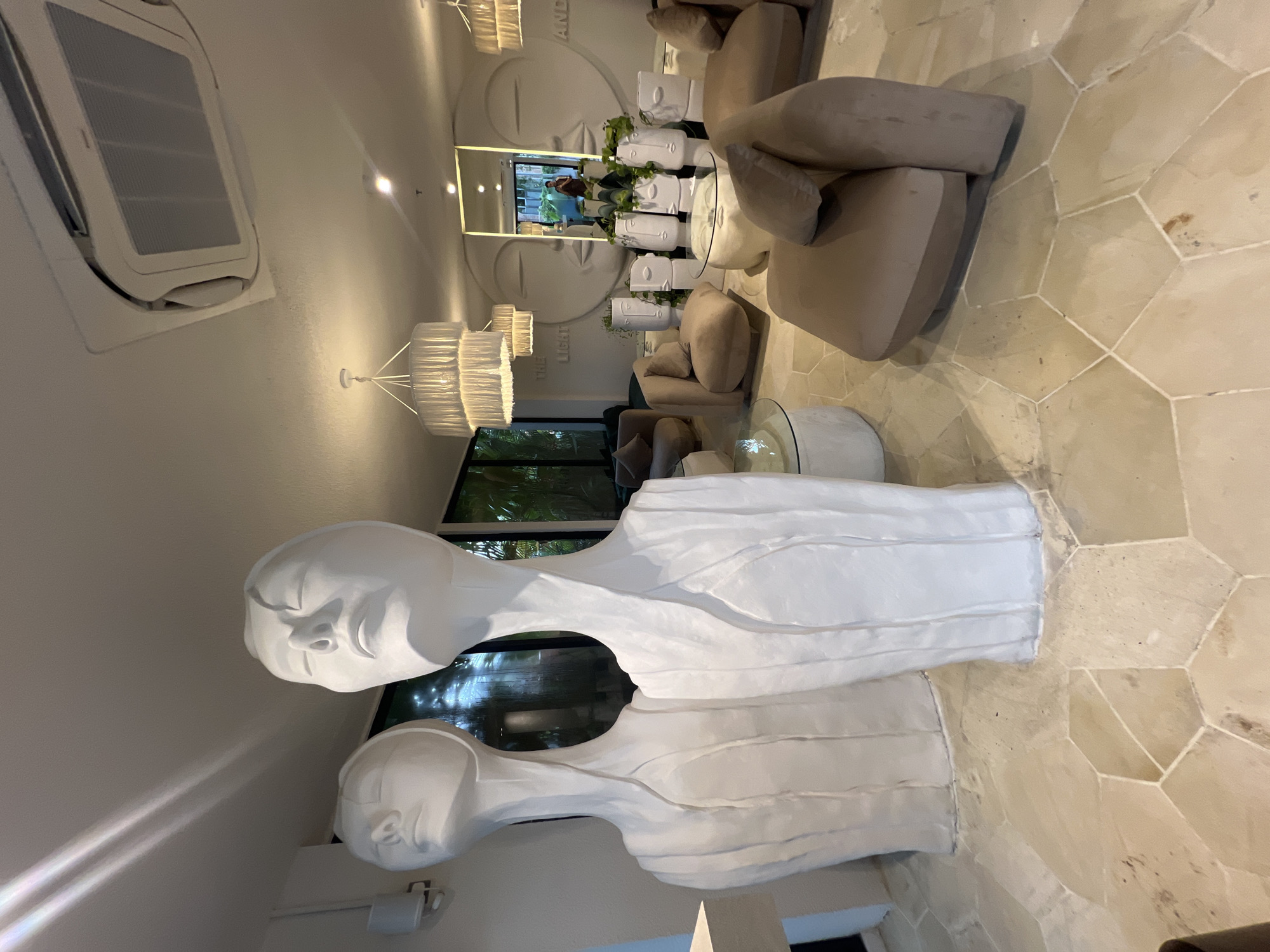 The image, taken in North Kuta, Indonesia, depicts an upscale, modern lounge or lobby area, likely within a hotel or resort. The scene is static, showcasing the room's elegant decor.
In the foreground, two prominent, white, abstract sculptures of human torsos and heads are visible. These sculptures have a textured, layered appearance, possibly made from a fabric-like or cast material, and stand on a light-colored, hexagonal tiled floor. The floor features varying shades of beige and tan tiles with visible grout lines, some showing minor scuffs or discoloration.
The mid-ground presents a comfortable seating area comprising plush, beige upholstered sofas and armchairs. Interspersed among the seating are several round coffee tables, some with glass tops. On one table, multiple white decorative planters, shaped like human heads, contain vibrant green plants, adding a natural touch.
The background wall features a large, artistic mural or relief depicting two stylized faces with closed eyes, conveying a sense of tranquility. Partially visible text on the wall reads "THE LIGHT" and "AND," suggesting part of a larger phrase. Above this, a large mirror reflects parts of the room, an outdoor area with green foliage, and a faint reflection of a person (likely the photographer).
The ceiling is adorned with large, multi-tiered white chandeliers featuring pleated fabric or paper shades, providing ambient lighting. A white air conditioning unit is visible on the upper left ceiling/wall. The overall ambiance is serene and sophisticated, blending contemporary design with artistic elements. The light suggests it is daytime, and the lush foliage implies a tropical setting. No specific activity is taking place; the room is set for relaxation or waiting.