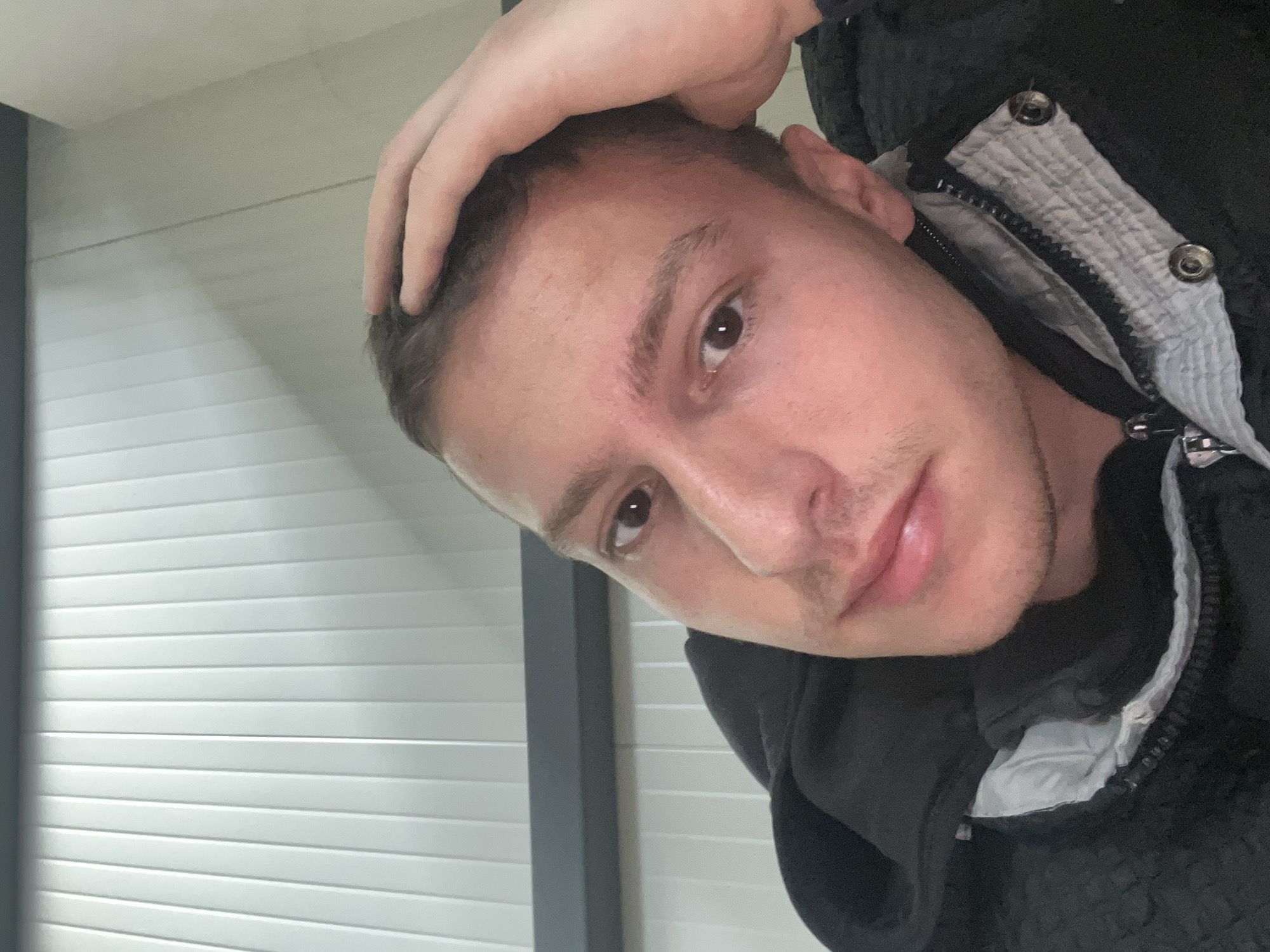 A man is shown in a close-up, tilted selfie. He is indoors, with white horizontal blinds visible in the background. He is light-skinned, with short brown hair, brown eyes, and stubble on his face. His hand is touching the top of his head. He is wearing a dark jacket with a grey and white trim and visible metal snaps. The location is unknown and cannot be determined from the image.