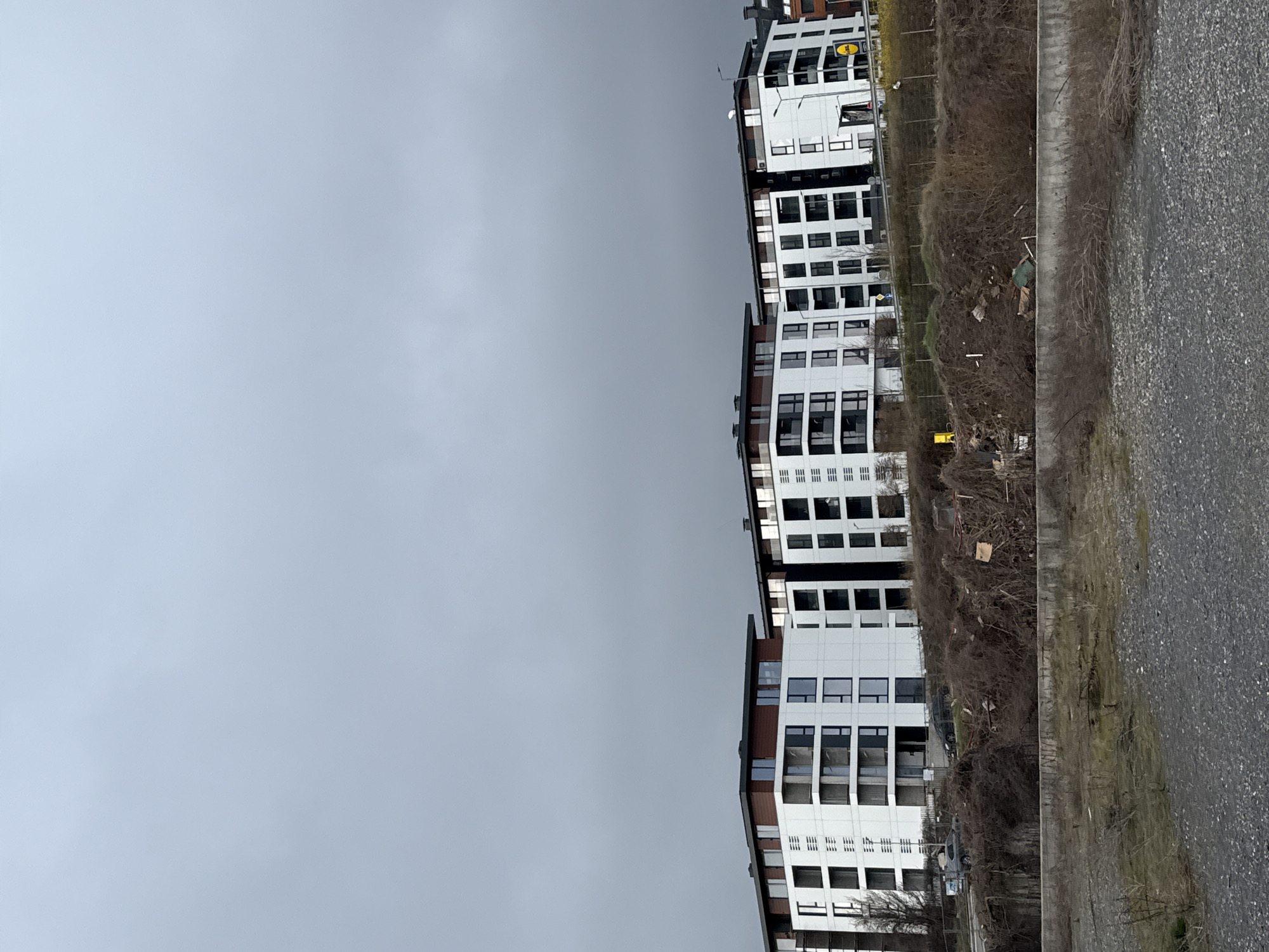 The image depicts a row of modern apartment buildings in Sofia, Bulgaria, seen on a cloudy day. The buildings are white with dark window frames and multiple stories, positioned adjacent to a concrete retaining wall and a paved area, possibly a road or sidewalk. Overgrown, bare vegetation separates the buildings from the walkway. The sky is overcast and gray. A few cars are seen near the buildings. A person stands near the vegetation.