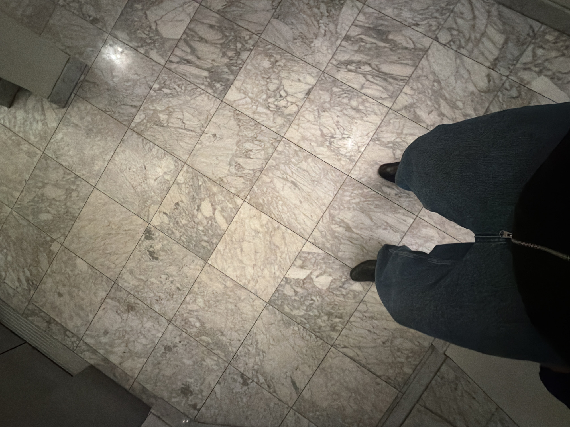 The image shows a portion of the floor, likely in a building in Paris, France, based on the marble tiling and the likely prevalence of such materials in Parisian architecture. The floor is covered with grey-white marble tiles arranged in a square grid pattern. There are some light reflections on the tiles. A person stands on the tiles, wearing dark shoes and blue jeans. The top portion of a dark jacket is visible. The person seems to be standing still. A portion of a wall or baseboard can be seen on the left side of the image.
