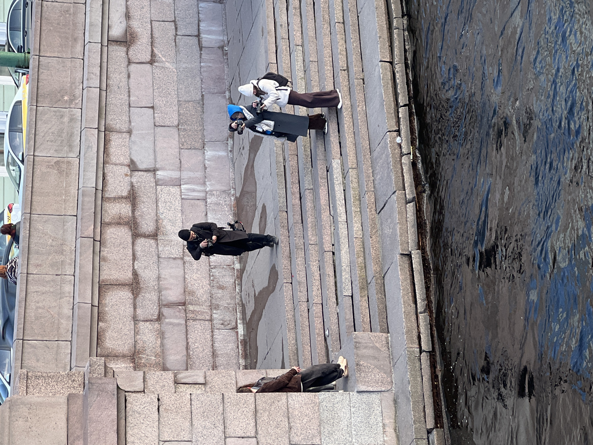 An eye-level aerial photograph shows a group of people on the granite embankment steps leading down to the Neva River in Saint Petersburg, Russia.
To the left of the steps, portions of several cars are visible.
On the steps, a person in brown lies prone at the bottom. Another person in black is higher up and is looking down, with what appears to be a camera. Further up the steps, two people, one wearing a blue hat and the other wearing a white hat, both appear to be taking photographs. The person in the blue hat is wearing a black coat; the person in the white hat is wearing a white jacket.
To the right of the steps, the Neva River is visible.