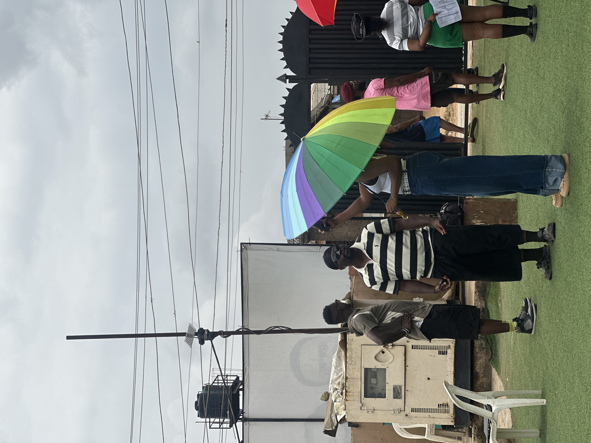 In Ashipa, Nigeria, a group of people are gathered outdoors on a cloudy day, standing on a patch of artificial green turf. A tall, dark metal pole with a solar panel mounted near its top stands prominently in the background, supporting overhead electrical wires. To the left of the pole is a large, white structure, possibly a building or a screen, with a black water tank on its roof. Adjacent to this structure is a beige generator. Several individuals are visible: one man in a gray t-shirt and black shorts stands with his hand on his hip, looking to the side. Another man in a black and white striped polo shirt and black shorts stands facing forward, wearing sunglasses and a black beanie. A person in the foreground holds a rainbow-colored umbrella over their head, partially obscuring their face. This person is wearing a white tank top and wide-leg blue jeans. Further in the background, a person in a pink t-shirt and dark shorts stands near a black spiked fence. To the right, a person in a gray and white striped shirt and a green skirt holds a piece of paper. Two white plastic chairs are positioned on the turf, one to the far left and another near the generator. The overall scene suggests a casual gathering or event taking place outdoors, possibly in a less developed area given the visible infrastructure and materials.