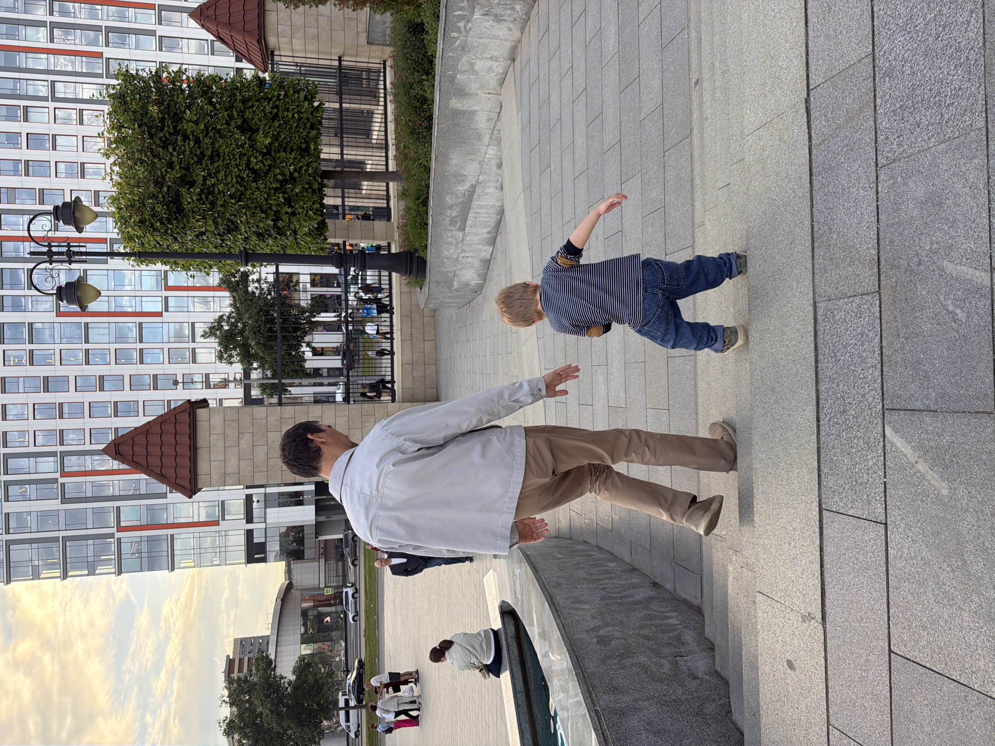 A man and a young boy are walking up a wide, light gray stone staircase in an urban outdoor setting. The man, from behind, is wearing a light beige jacket and tan pants. The boy, also from behind, has blond hair and is wearing a striped long-sleeved shirt with blue jeans. They are walking away from the viewer, towards a tall, modern building with a white facade and red accents. The building has many rectangular windows. In the background, there are trees, a lamppost with two lights, a metal fence, and other people walking. The staircase is made of large, rectangular stone slabs with visible grout lines. The lighting suggests it is late afternoon or early evening.