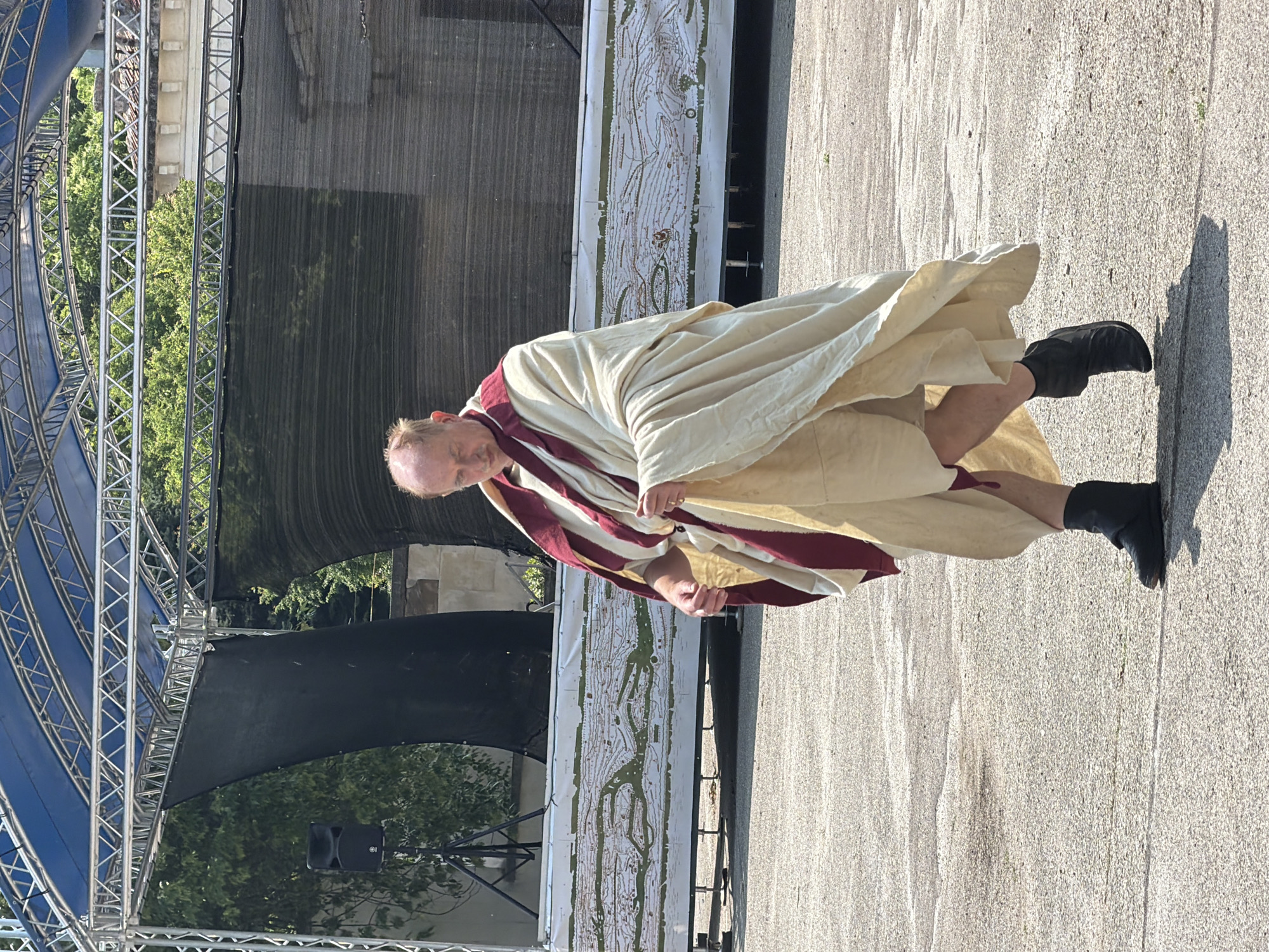 An older man, partially balding with light-colored hair on the sides, walks from left to right across a light gray, textured asphalt or concrete surface. He is dressed in a cream-colored, toga-like garment draped over his body, with a deep red or maroon stole over his left shoulder and chest. His lower legs are visible, and he wears black, boot-like footwear that covers his ankles. He looks downwards, and his hands appear to be adjusting his garment. His left foot is lifted mid-stride.
In the background, an elevated outdoor stage structure is present. It features a blue fabric canopy stretched over a silver metal truss framework. Dark, possibly black, mesh or fabric screens hang from the truss, serving as a backdrop. A large, white banner or board runs along the lower front edge of the stage, displaying a detailed, predominantly green and white topographical map-like illustration, possibly depicting historical terrain or battle movements. To the far left, a black speaker on a tripod stand is visible next to the stage. Green trees and dense foliage are visible behind and to the sides of the stage structure. To the right, parts of a light-colored building with a tiled roof are discernible through the trees and gaps in the stage backdrop. The scene is illuminated by bright daylight, casting visible shadows on the ground.
Based on the distinctive topographical map on the stage banner, which likely represents historical events, the location is an outdoor venue in Pleven, Bulgaria, likely associated with historical or cultural events related to the Pleven Epopee 1877, potentially in or near Skobelev Park or the Panorama "Pleven Epopee 1877" complex.