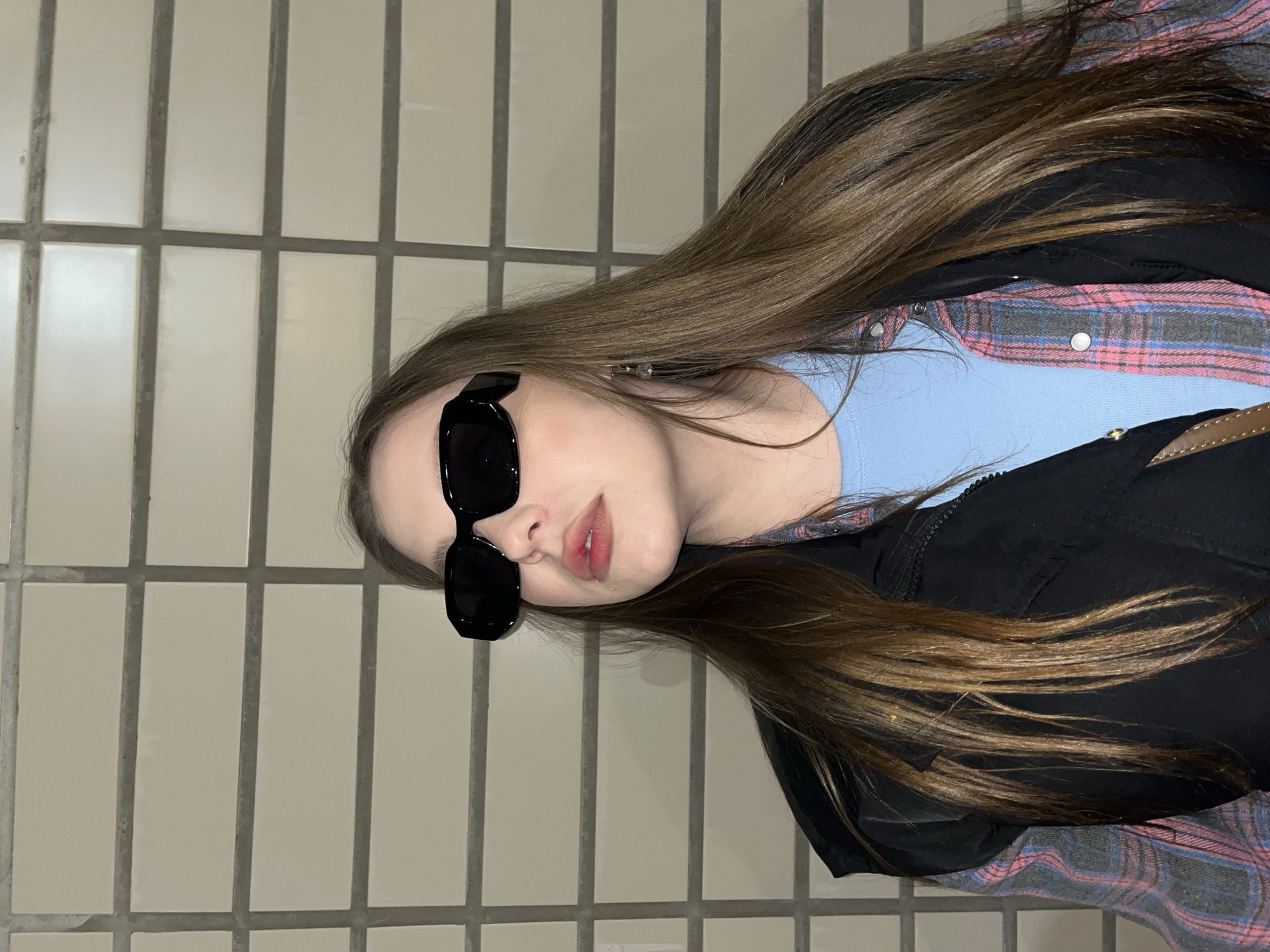 A portrait-style photograph, rotated ninety degrees clockwise, features a young woman posing indoors against a light-tiled wall. She has long, straight, medium brown hair. Her eyes are obscured by black, rectangular sunglasses. Her lips are slightly parted, revealing a subtle pink hue. She is wearing a light blue top underneath an unbuttoned pink, blue, and gray plaid shirt. Over these layers, she wears a black jacket or vest, with a brown strap, possibly from a bag, visible on her right shoulder. The background consists of uniform, light-colored rectangular tiles with dark gray grout lines. No specific activity beyond posing is evident. No text is visible in the image. The location context provided is Kudrovo, Russia.