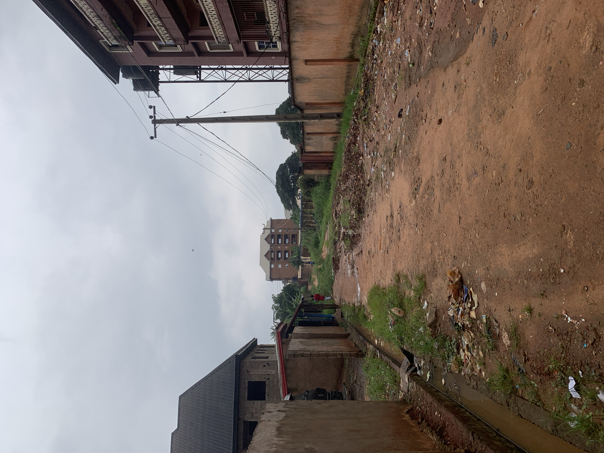 The image depicts a narrow, unpaved dirt road in Nkpor, Nigeria, lined with a mixture of residential buildings and what appear to be informal structures. The road surface is uneven and shows signs of recent rainfall with muddy patches and puddles. Litter is visible along the sides of the road and in the immediate foreground, including plastic bags, paper, and other debris.
On the left, a partially constructed building with a dark, sloped roof and unplastered brick walls is prominent. Adjacent to it is a low-lying structure with a reddish roof, possibly a small shop or dwelling. A dark-colored vehicle is partially visible behind a concrete wall in front of this structure. A drainage channel runs along the edge of the property, also containing some refuse.
On the right side of the road, a multi-story building with a reddish-brown facade and balconies with white railings stands tall. A wooden utility pole with electrical wires runs alongside this building. Further down the road, another multi-story building with a similar color scheme is visible in the distance, along with lush green trees.
A single individual, wearing a red garment, is walking away from the camera on the dirt road, heading towards the background. Another person, less distinctly visible, is further down the road. The sky is overcast and grey, suggesting a cloudy or potentially rainy day. The overall impression is one of a developing urban environment with evident challenges related to infrastructure and waste management.