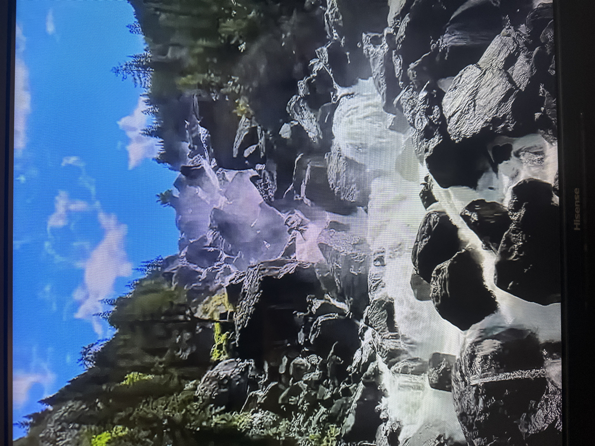 The image depicts a powerful waterfall cascading down a rocky, mountainous slope. Lush green trees are visible on the higher slopes, contrasting with the dark, rugged rocks that form the waterfall's path. The sky above is a bright blue with scattered white clouds. The water appears frothy and turbulent as it rushes over the rocks, creating a misty atmosphere in the middle section of the waterfall. The scene is dominated by natural elements: rock, water, and vegetation. There is no indication of the city of Kyiv, Ukraine, or any urban features in this image.