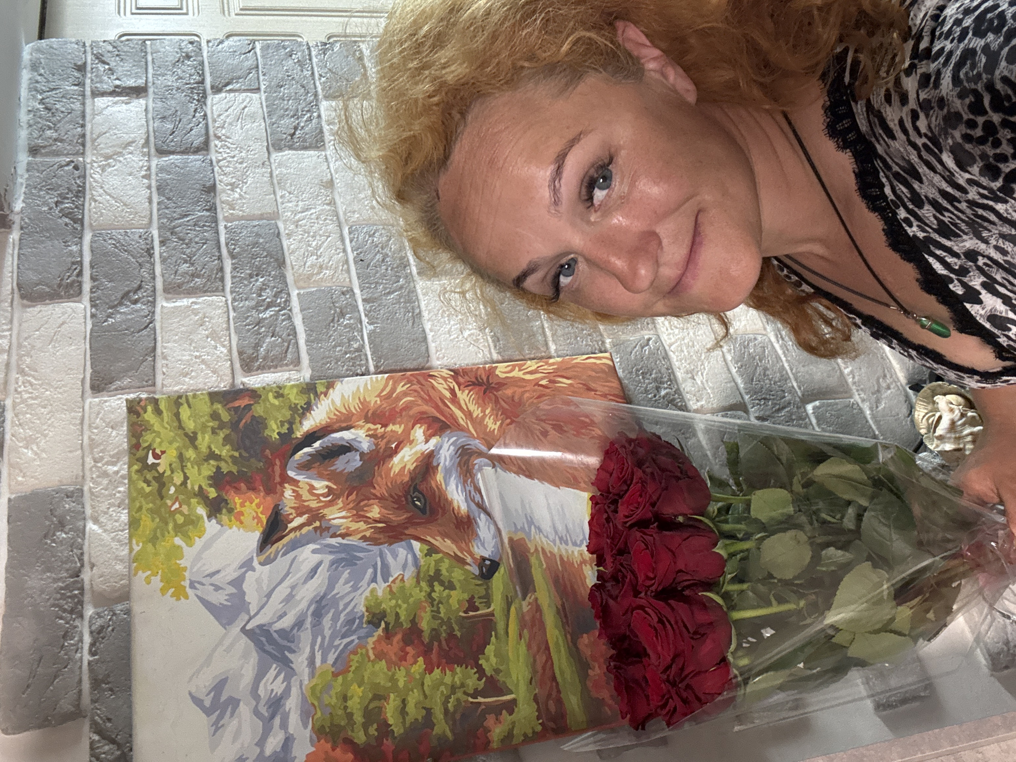 In Chaikovsky, Russia, a woman with curly blonde hair and blue eyes stands in front of a wall with a grey and white brick pattern. She is holding a bouquet of red roses wrapped in clear plastic. To her left, a painting of a fox in a natural landscape hangs on the wall. The woman is wearing a black and white leopard-print top with black lace trim and a necklace with a green pendant. The overall lighting suggests an indoor setting, possibly a home or a gift shop.