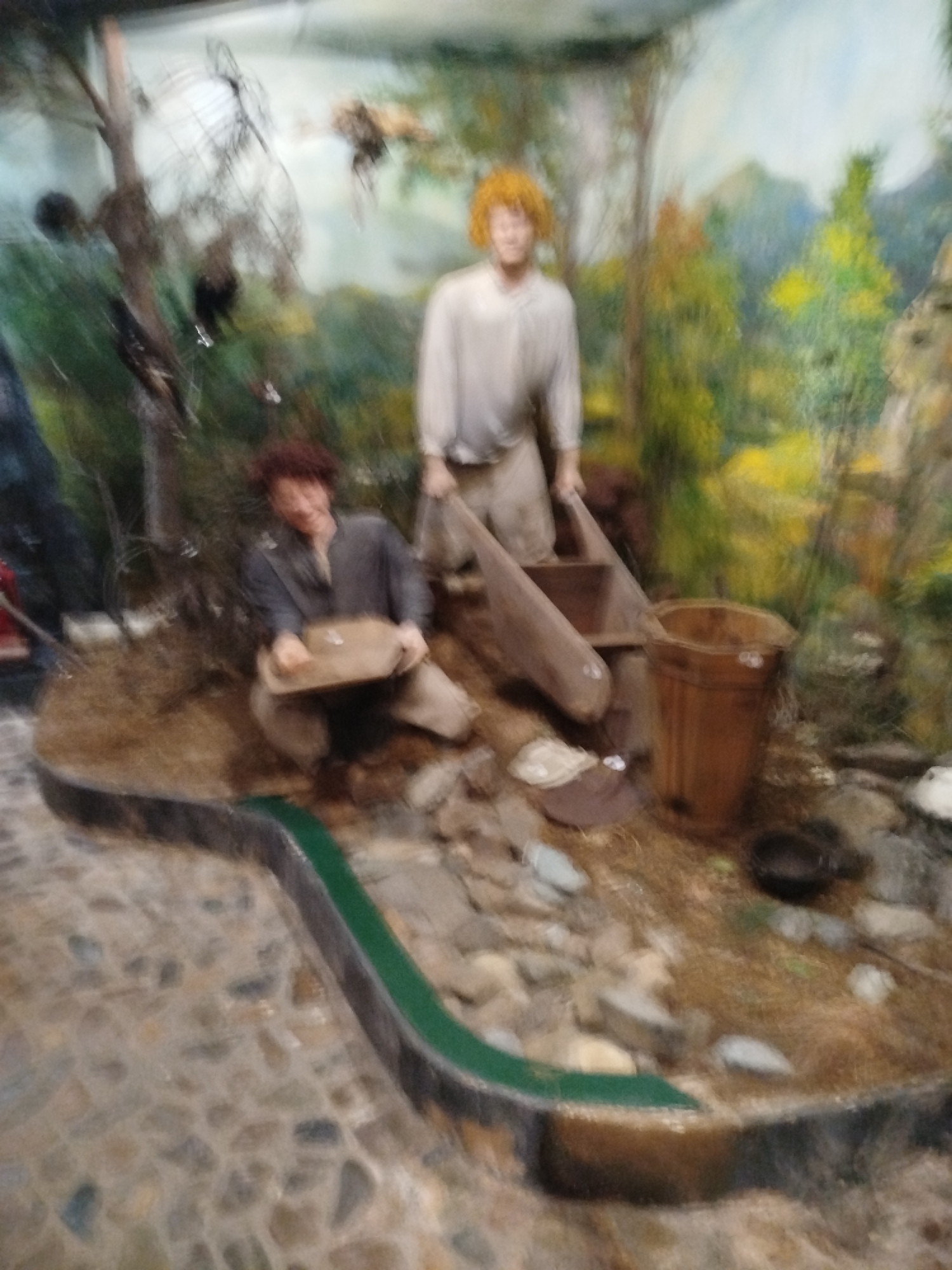 The image depicts a diorama of gold panning activity, likely displayed in a museum. Two male mannequins are present. One, positioned on the right, has bright orange hair and is wearing a light-colored shirt and trousers. He is standing and appears to be holding a wooden sluice box. To the left, a second mannequin with dark, curly hair is seated and is wearing a dark shirt and trousers. He is holding a wooden pan filled with what appears to be dirt or gravel. The diorama is set against a painted backdrop of a natural landscape with trees and mountains. The foreground is a simulated stream bed with rocks and what looks like a trough with a green liner. A wooden bucket is also visible. The overall scene suggests a historical or educational exhibit related to gold mining. The city of Tashtagol, Russia, is known for its mining industry, making such a diorama plausible for a local museum.