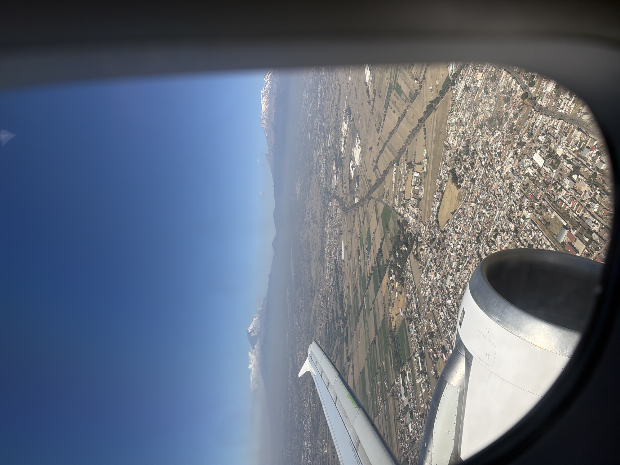 An aerial photograph, taken from an airplane passenger window, depicts the landscape above the city of Santa Ana Xalmimilulco, Mexico, in the state of Puebla. The image frames a portion of a white aircraft wing with green detailing on its leading edge in the upper left, and a segment of a white and silver turbofan engine nacelle in the lower left, both partially obscured by the dark, curved window casing. Below the aircraft, the ground reveals a dense urban area, characterized by numerous light-colored, closely packed buildings, which corresponds to Santa Ana Xalmimilulco and its adjacent settlements. This urban development is interspersed with and transitions into expansive agricultural fields, organized into distinct rectangular and irregular plots of varying hues, indicating different cultivation or fallow states. In the distant background, under a clear blue sky with some atmospheric haze, two prominent snow-capped volcanoes are visible: Iztaccíhuatl to the left, partially covered by clouds near its summit, and Popocatépetl to the right, displaying a more defined snow cap. The entire scene is brightly illuminated by daylight. No individuals, specific actions, or direct interactions are visible.