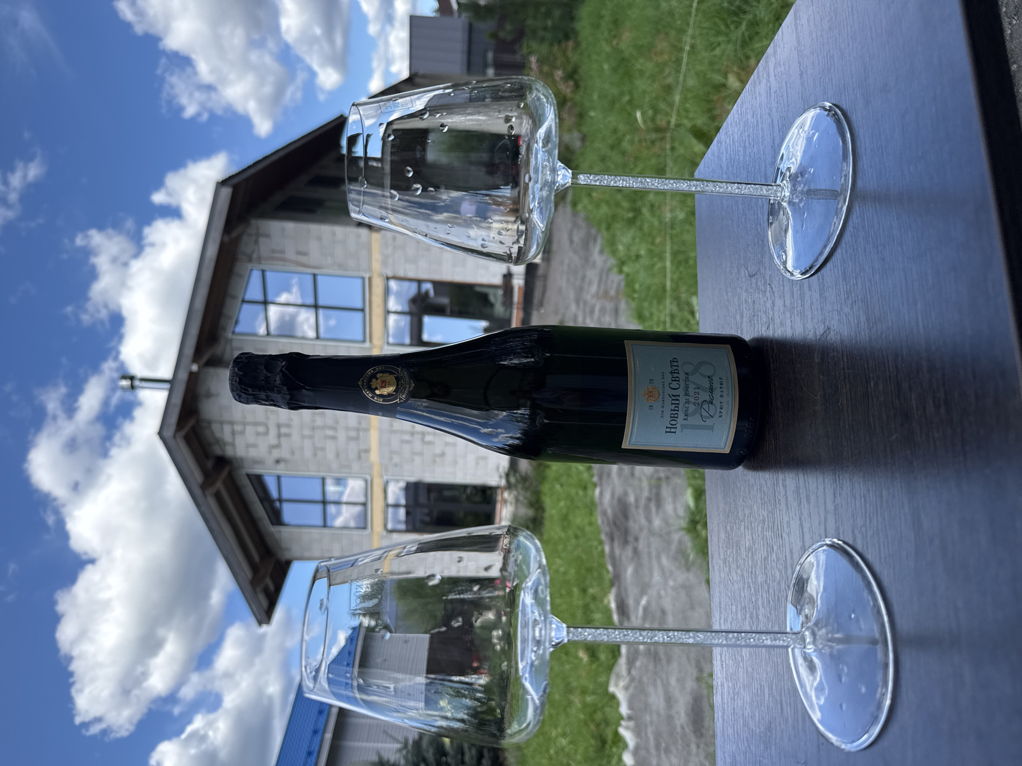 On a dark wooden table, outdoors, a bottle of sparkling wine and two empty, stemmed wine glasses are positioned. The bottle, dark green glass with a black foil capsule, is upright, positioned centrally. It has a light blue label with gold lettering. The wine glasses are clear, with the left glass containing condensation on the interior and the right glass exhibiting similar condensation. Their stems are clear and appear to be textured or filled with small, reflective particles.
In the background, a house under construction is visible. It is a large, modern structure with light gray brick walls and dark-framed windows. The roof is dark gray with exposed wooden rafters. A metal chimney extends from the roof. To the left of this structure, a smaller house with a bright blue roof is partially visible.
The environment is grassy with some patches of gray gravel or pavement. The sky is bright blue with scattered white, puffy clouds. The overall setting suggests a suburban or rural area. The location is identified as Steklyannyi, Russia.