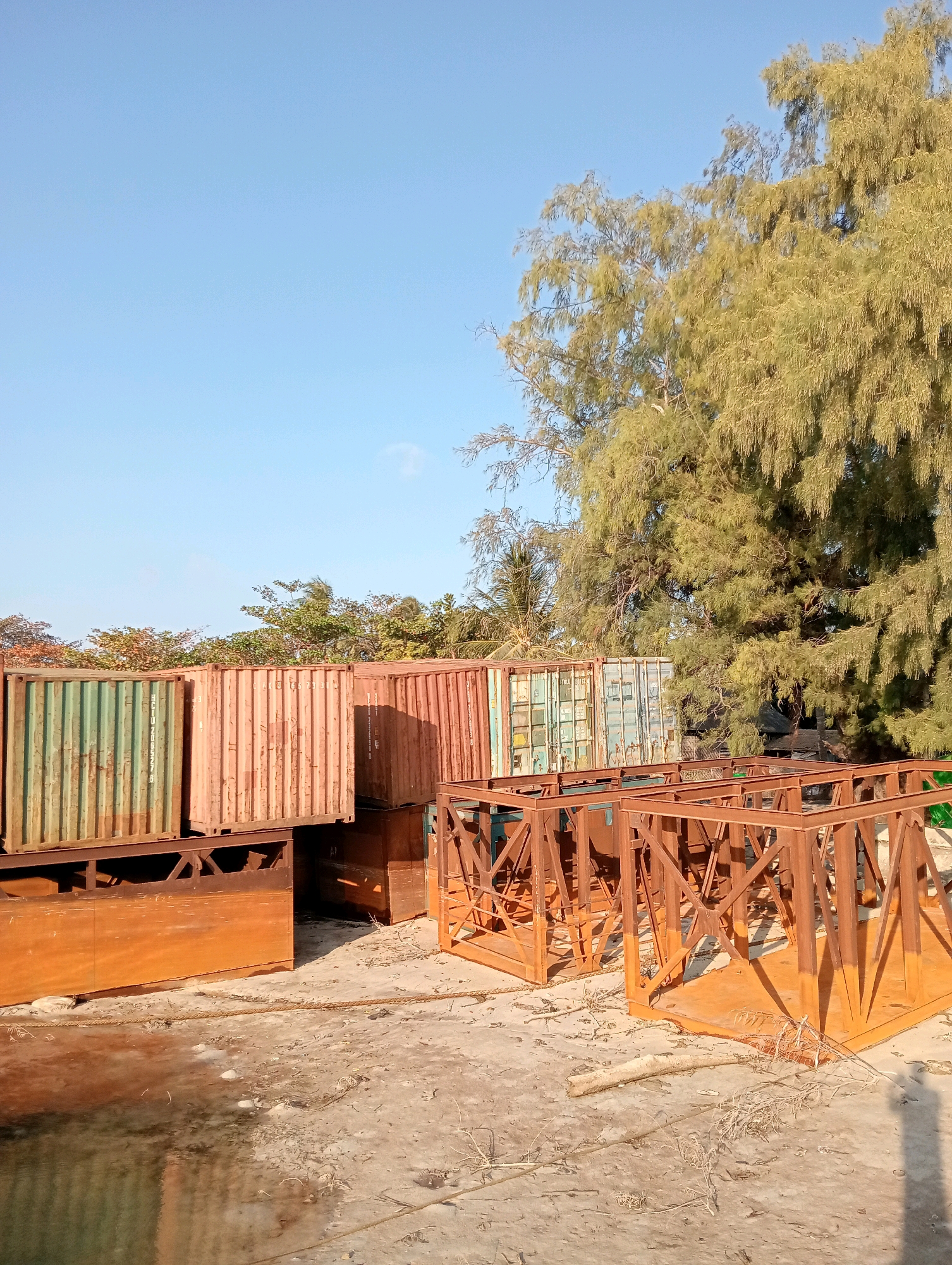 In Pagerungan Kecil, Indonesia, an outdoor scene depicts several stacked shipping containers. The containers, some appearing weathered and slightly rusted, are arranged in rows. In the foreground, to the right, are several metallic, rust-colored structures that appear to be part of a framework or support system, possibly related to the shipping containers. The ground is sandy and uneven, with sparse dry vegetation. Behind the containers and framework, a large tree with dense, feathery foliage dominates the right side of the image, with other trees and foliage visible in the background. The sky is clear and blue. The scene appears to be a storage or staging area. There are no people visible in the image.