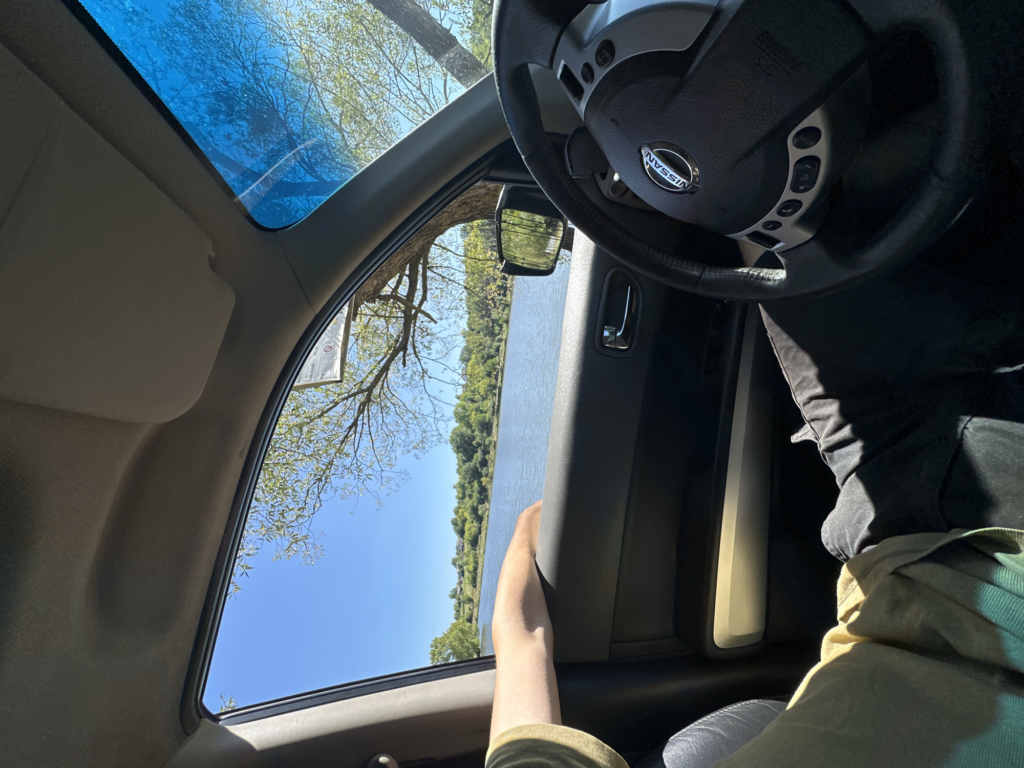 The image shows the interior of a vehicle with a person's arm resting on the open car door. Through the open window, a body of water, trees, and a clear blue sky are visible. The steering wheel of the car, with the Nissan logo, is in the foreground. The scene appears to be a lakeside setting on a sunny day. It is impossible to determine the location in the city of Makiv, Ukraine from the visual information provided in the image.