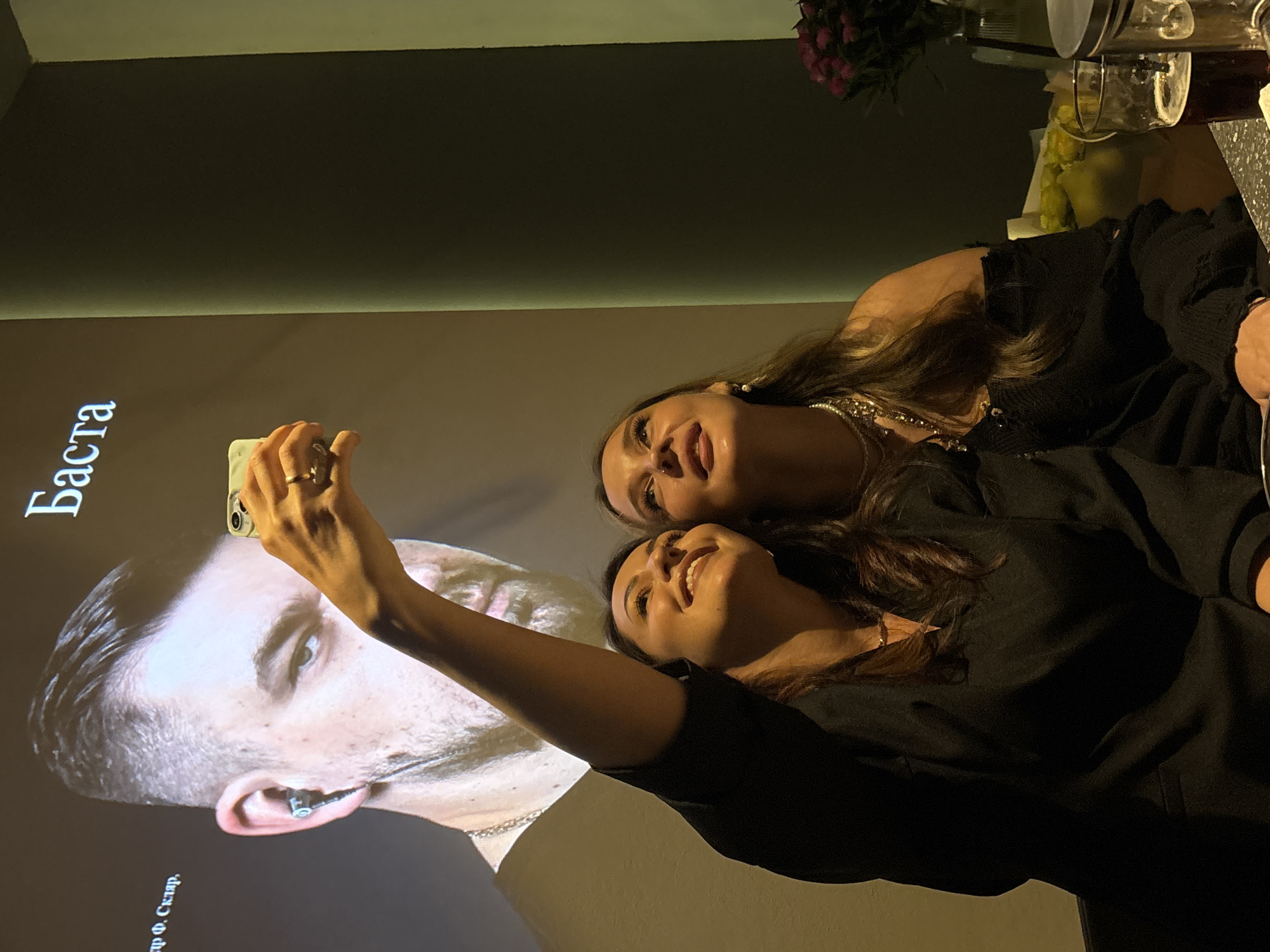 Two women are captured in a selfie in a dimly lit indoor setting, likely a restaurant, bar, or event venue in Chaikovsky, Russia, during the evening or night.
In the foreground, a woman with long, dark hair holds a light-colored smartphone, taking the selfie. She smiles, wears a black top, and has a ring on her left ring finger. Behind her, another woman with long, dark hair and a black top smiles broadly, showing her teeth. She wears several pearl necklaces and has a small nose piercing. Both women appear joyful and are looking towards the camera.
A large projection of a man's head is displayed on the wall behind them. The man has short, dark hair, a beard, and an ear piercing. Cyrillic text on the projection reads "Баста" (Basta), and below the man's head, "гр. Ф. Скляр" (gr. F. Sklyar) is visible. These are names of Russian musicians, suggesting a music-related event or venue.
To the far right, partially visible background elements include a floral arrangement with pink flowers and a glass pitcher or decanter with a dark liquid on a table. The overall mood conveyed is cheerful and social.