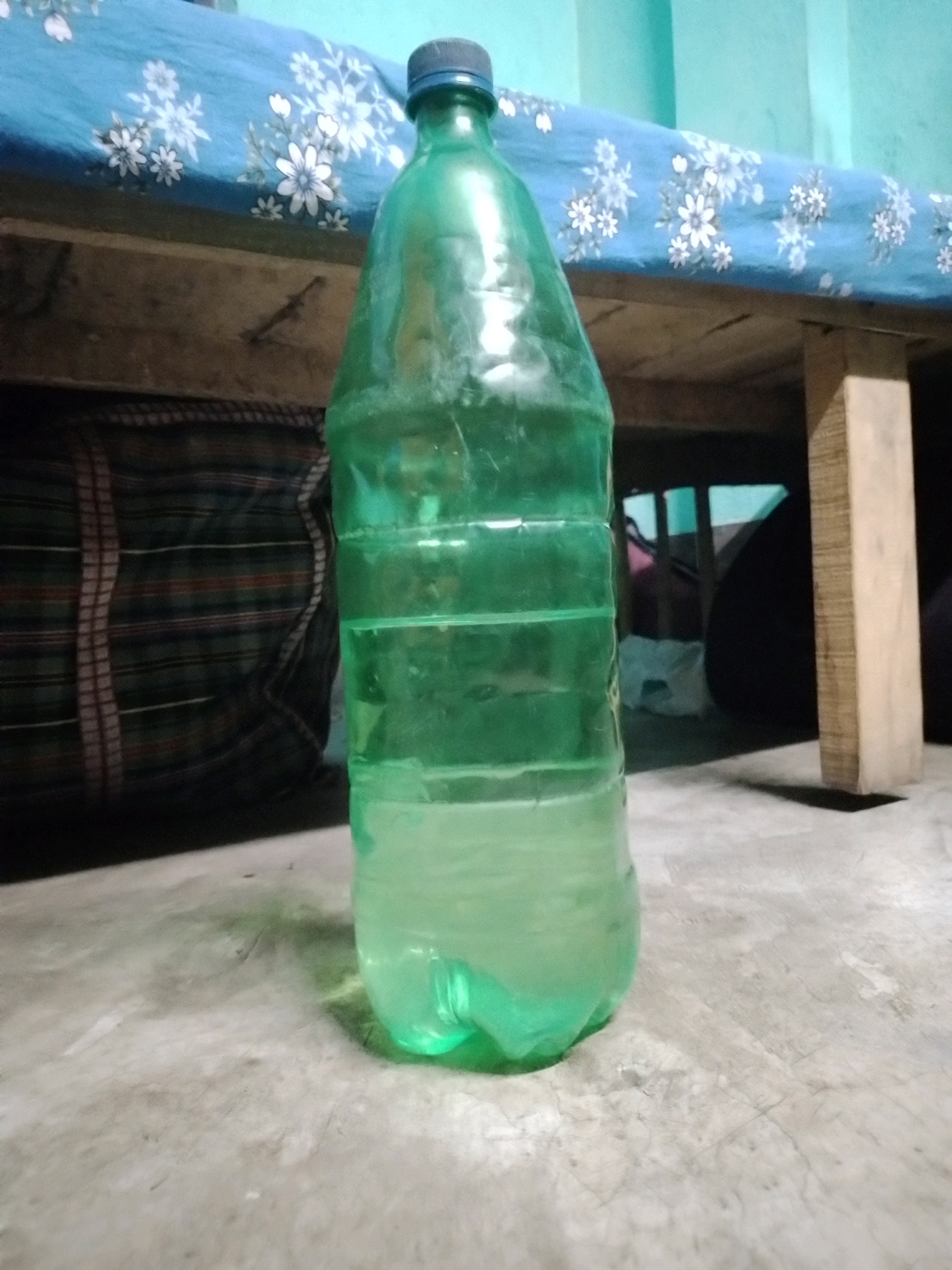 A green plastic bottle filled with a pale green liquid stands on a concrete floor. The bottle is centrally positioned in the foreground, with its cap visible at the top. The bottle appears to be half-full, with a distinct line separating the liquid from the air above it. In the background, a wooden structure, possibly part of a bed frame, is visible. A blue cloth with a white floral pattern is draped over the wooden structure. To the left of the bottle, a fabric with horizontal stripes in various colors is partially visible. The environment suggests an indoor setting, likely a room. The lighting is dim, casting shadows on the concrete floor. The overall scene is devoid of any human presence or activity. The image was taken in the city of South Dumdum, India.