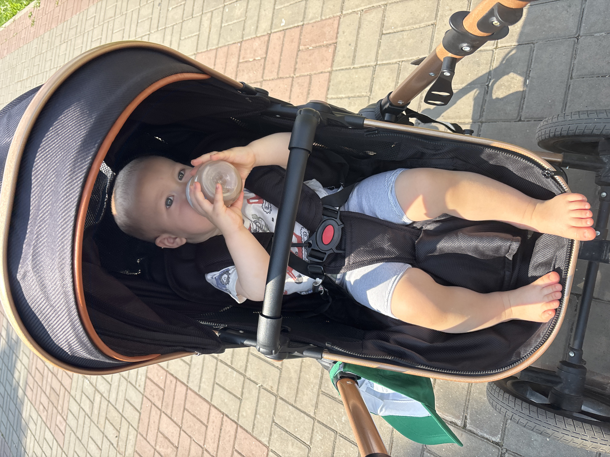 A light-skinned baby sits in a stroller on a sidewalk. The baby is drinking from a clear bottle. The stroller has a black fabric interior with brown trim. The baby is wearing a white t-shirt with a graphic design and grey shorts. The sidewalk is made of interlocking bricks in various shades of red and tan. The sun is casting shadows on the sidewalk. The location is the city of Naberezhnye Chelny, Russia.
