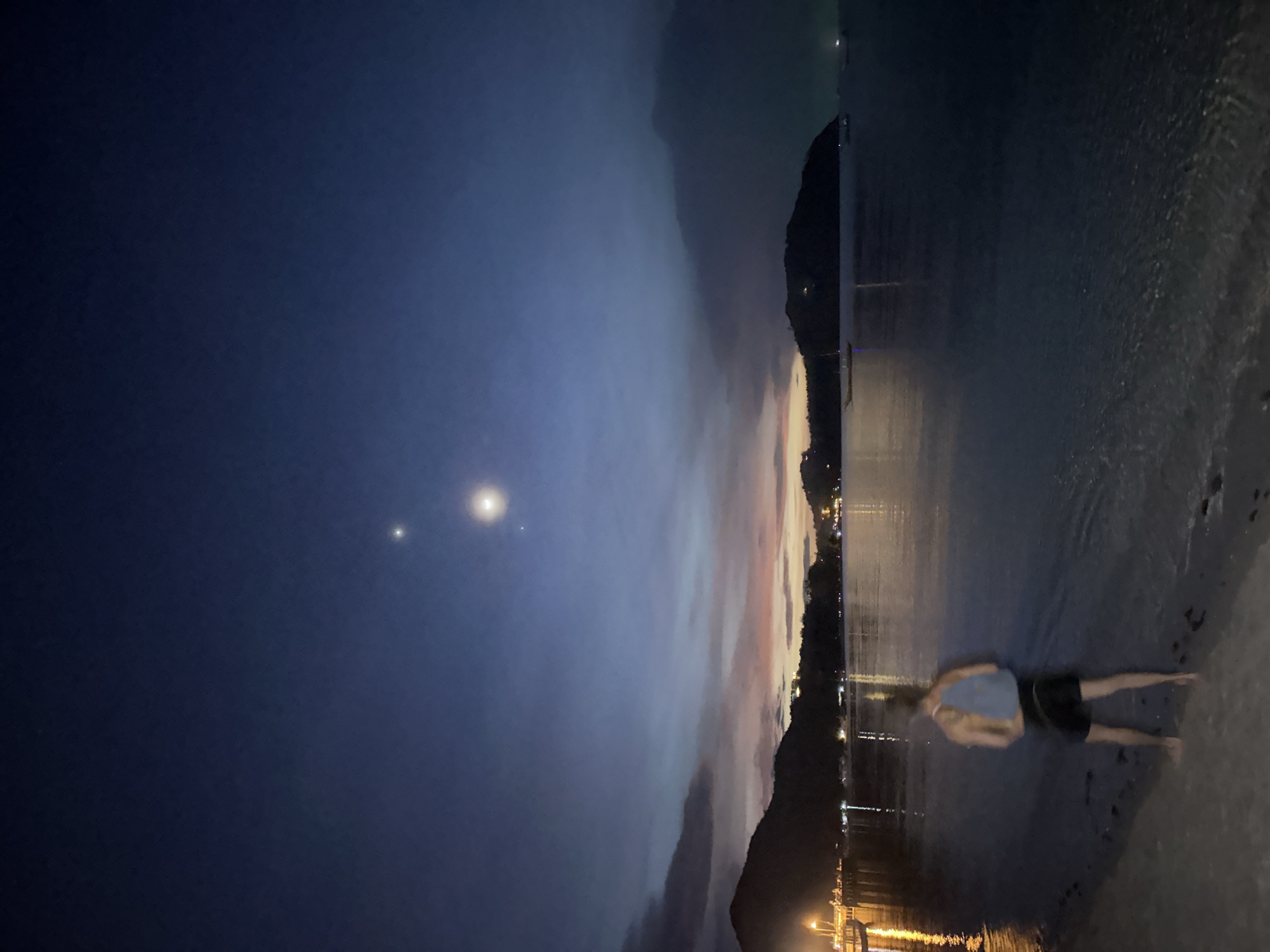 A person is captured in a full shot, lying face down on a wet sandy beach. They are wearing a light blue tank top and black shorts. The water laps gently at the shore, reflecting the faint lights from a distant town that lines the base of a dark, silhouetted mountain. The sky above is a deep, twilight blue, with two bright celestial bodies visible; one is a prominent, large circular light, and the other is a smaller, fainter point of light nearby. Wisps of clouds streak across the sky, catching the last vestiges of sunset with hues of pink and orange near the horizon. The overall scene evokes a sense of tranquility and calm, as if the person is enjoying a peaceful evening by the sea. The image appears to have been taken at dusk or early night.
