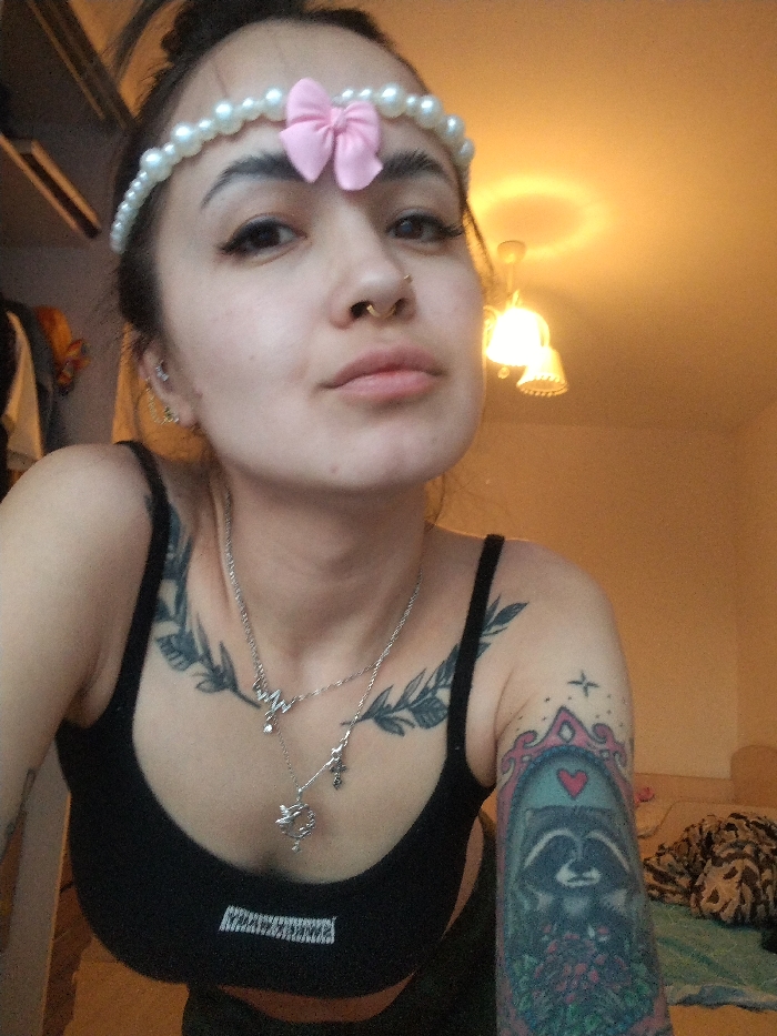 This image depicts a young adult female subject positioned centrally in a tight indoor shot, presumed to be within a residence in Noviy, Russia. The subject faces the camera directly, her head slightly tilted. She has dark hair styled back, wearing a white pearl-like headband adorned with a small pink fabric bow on her forehead. Her facial features include visible eyeliner and a septum piercing. Multiple small piercings are visible on her left ear.
She wears a black tank top or sports bra, revealing tattoos on her upper chest and left arm. A vine or leafy pattern tattoo extends across her upper chest. On her left arm, a sleeve tattoo features a framed image of a raccoon holding a bouquet, with a pink heart above its head and small stars above the frame. Two silver-colored necklaces are worn; one depicts a heartbeat line design with a small pendant, and the other is a longer chain with various charms, including a small cross and a more ornate, larger pendant.
The background is dimly lit with a warm, yellowish artificial light source, identified as a multi-bulb ceiling fixture with bell-shaped shades visible in the upper right. To the left, a partial view of an open closet or wardrobe with hanging garments is discernible. To the far right, out of focus, what appears to be a bed with patterned bedding is present. The walls are a light, non-distinct color, illuminated by the warm interior lighting. No active interaction beyond posing for the photograph is depicted.