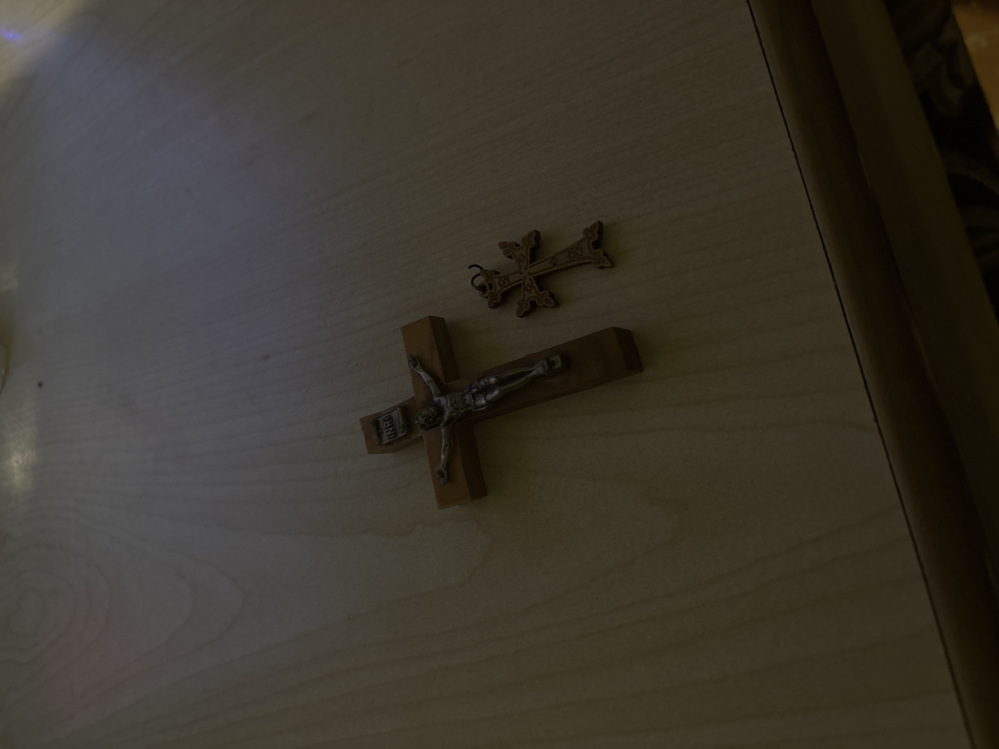 The image shows two crosses on a light-colored wooden surface, likely a table or desk. The larger cross is a traditional crucifix with a metal figure of Jesus. The smaller cross has a decorative design and an eyelet suggesting it could be worn as a pendant. The lighting is somewhat dim. The image does not contain any information about the location being Yerevan, Armenia, or any other specific location.