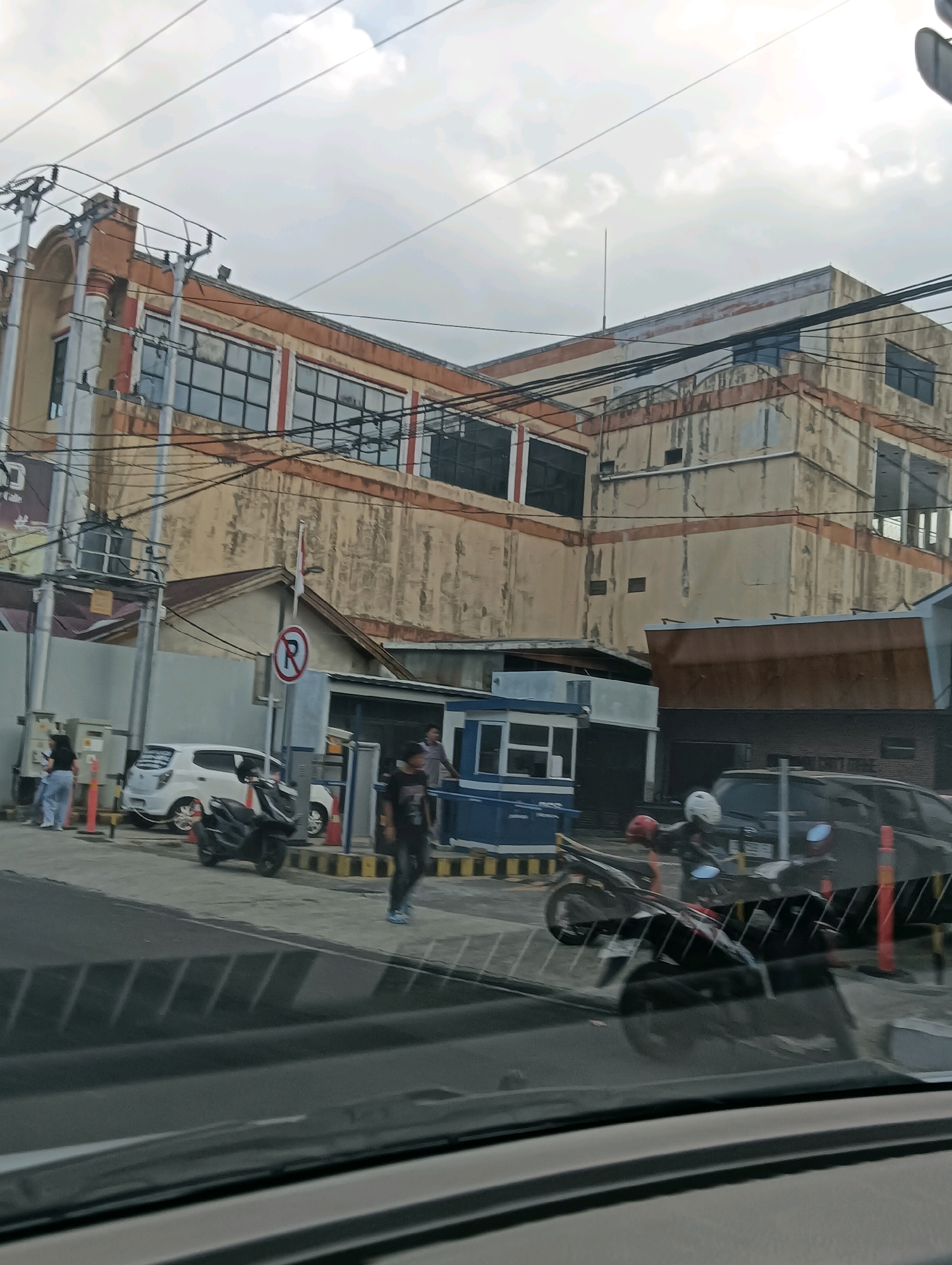 The image captures a street scene in Tomohon, Indonesia, featuring a multi-story building with a weathered facade and orange and beige accents. The building has numerous windows, some of which appear boarded up or dark. Overhead, a network of power lines crisscrosses the sky, which is overcast.
In the foreground, a dark-colored car is partially visible, reflecting the surrounding environment in its windshield. To the right of the car, a black motorcycle with a white helmet is parked. Further back, a smaller white car is parked. A black motorcycle is also parked behind the white car.
Several people are present. Two individuals stand near the white car, and another person is walking in the middle ground. Two more individuals are visible near a blue and white booth-like structure. A red and white "P" in a circle sign, indicating parking, is also visible. The ground appears to be paved, with some yellow and black striped barriers suggesting a defined area. The overall impression is that of a commercial or mixed-use urban environment.