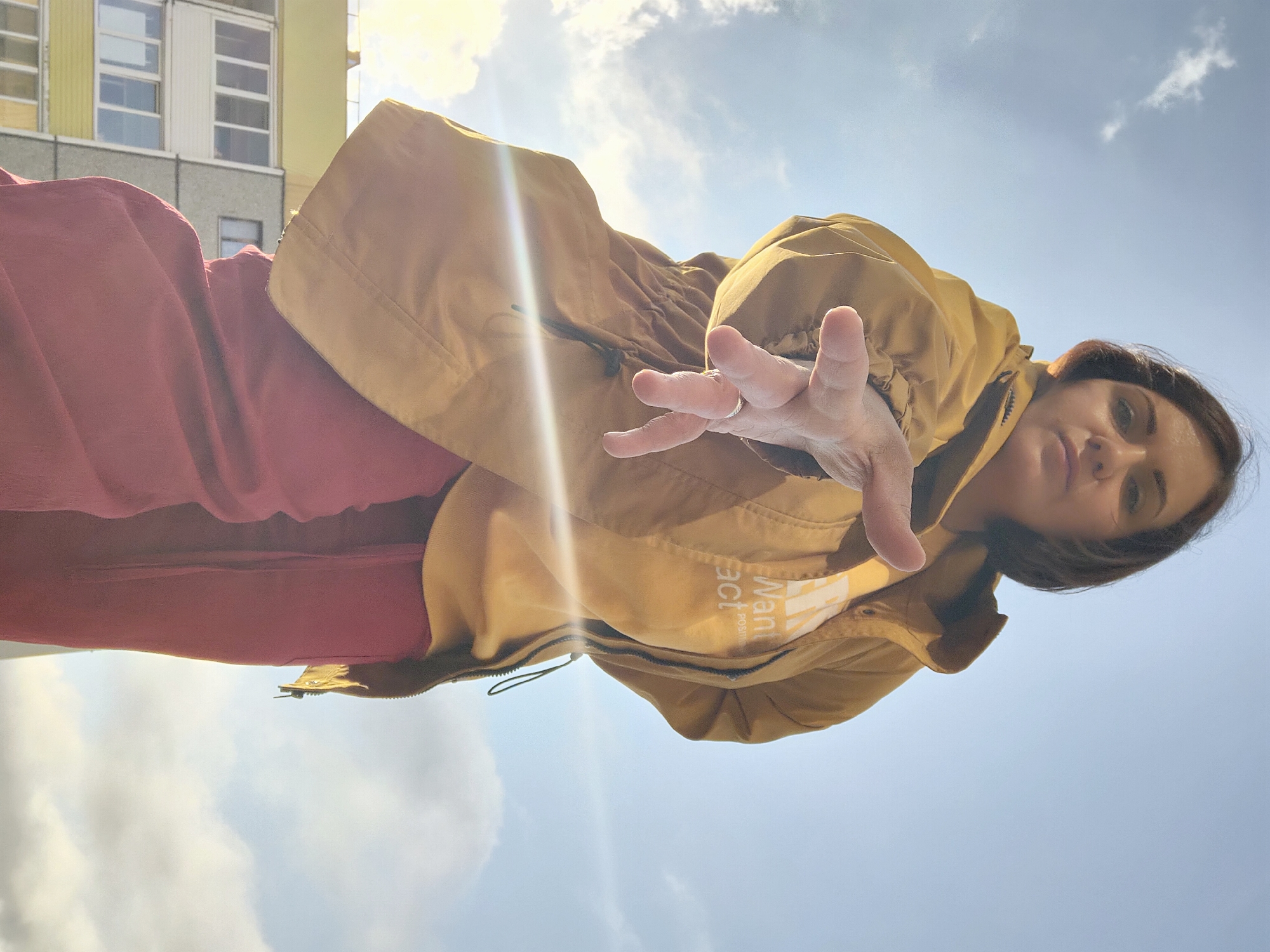 The image shows a woman in Марковский, Russia, from a low-angle perspective. She is wearing a mustard-colored jacket with the word "Want" visible on it and red pants. She is extending her hand towards the camera. In the background, there is a building with white windows and a light-colored facade, and the sky is visible with some clouds. The sun is shining, creating a bright glare.