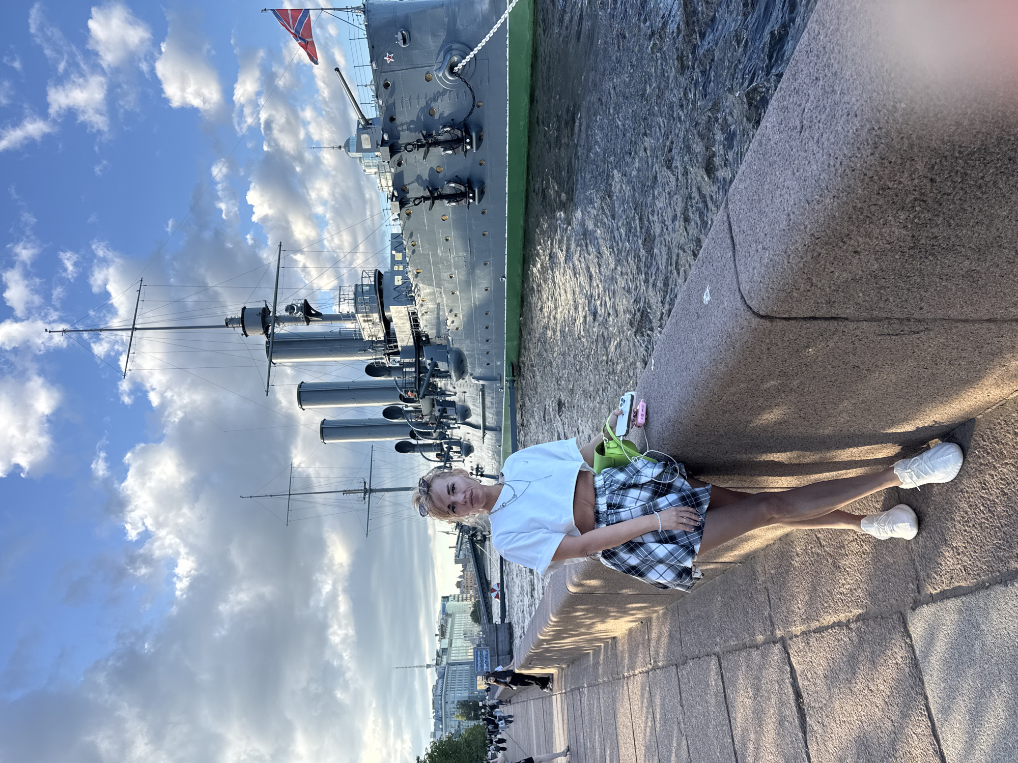 A woman is standing on a stone embankment in front of a moored warship in Saint Petersburg, Russia. The woman is wearing a white crop top, a plaid skirt, and white sneakers. She is holding a phone and a bright green bag. In the background, the warship, identified as the cruiser Aurora, is visible with its cannons and masts. The ship is flying a Russian naval flag. The water is choppy, and the sky is partly cloudy with dramatic light. In the distance, buildings of Saint Petersburg can be seen along the waterfront.
