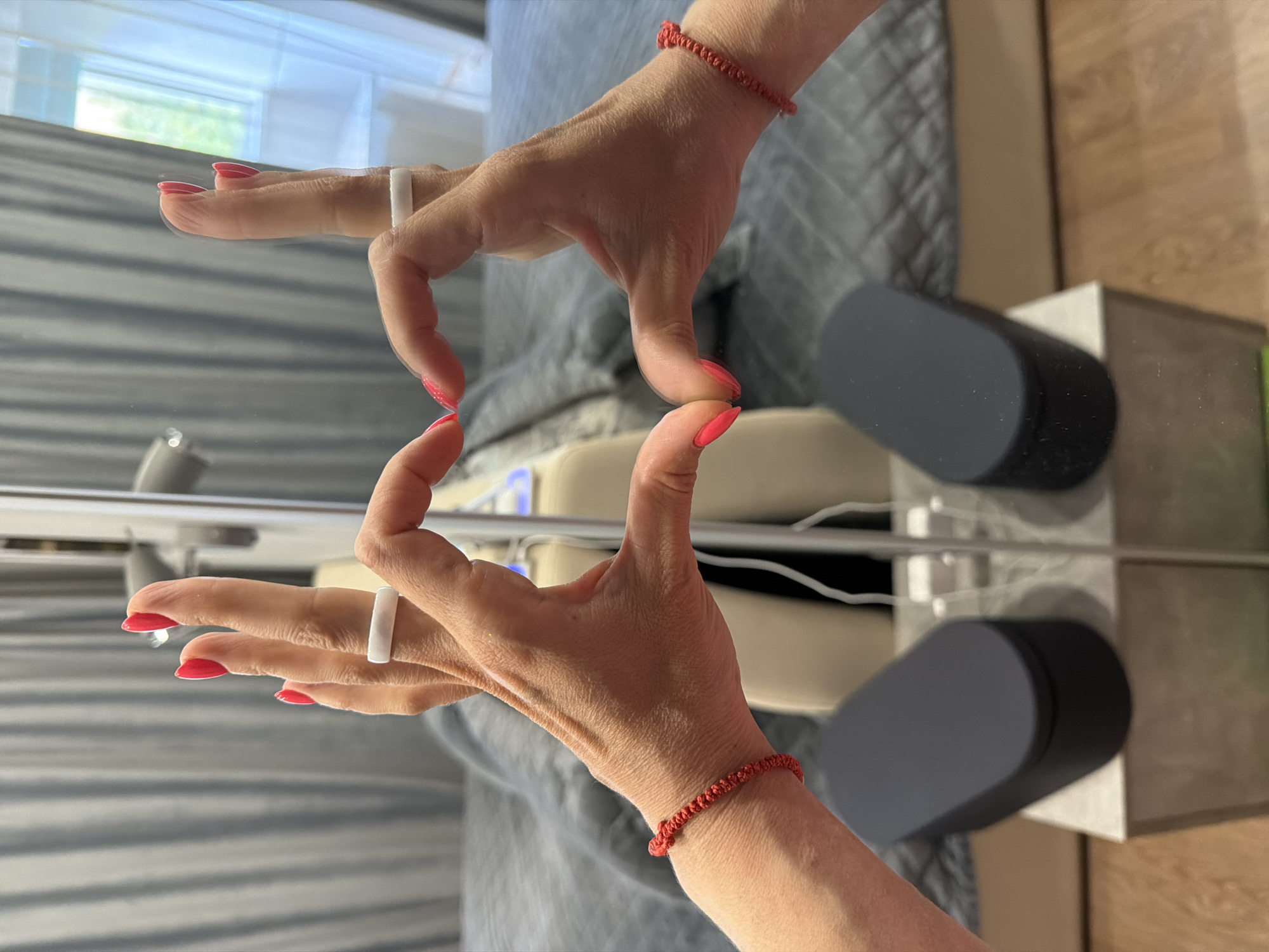 The image depicts two hands forming a "Z" shape, likely in front of a mirror. The hands have red nail polish and one hand wears a white ring, while the other has a red string bracelet. The background appears to be a bedroom with a bed and window. There is no evidence to suggest this image was taken in Birobidzhan, Russia.
