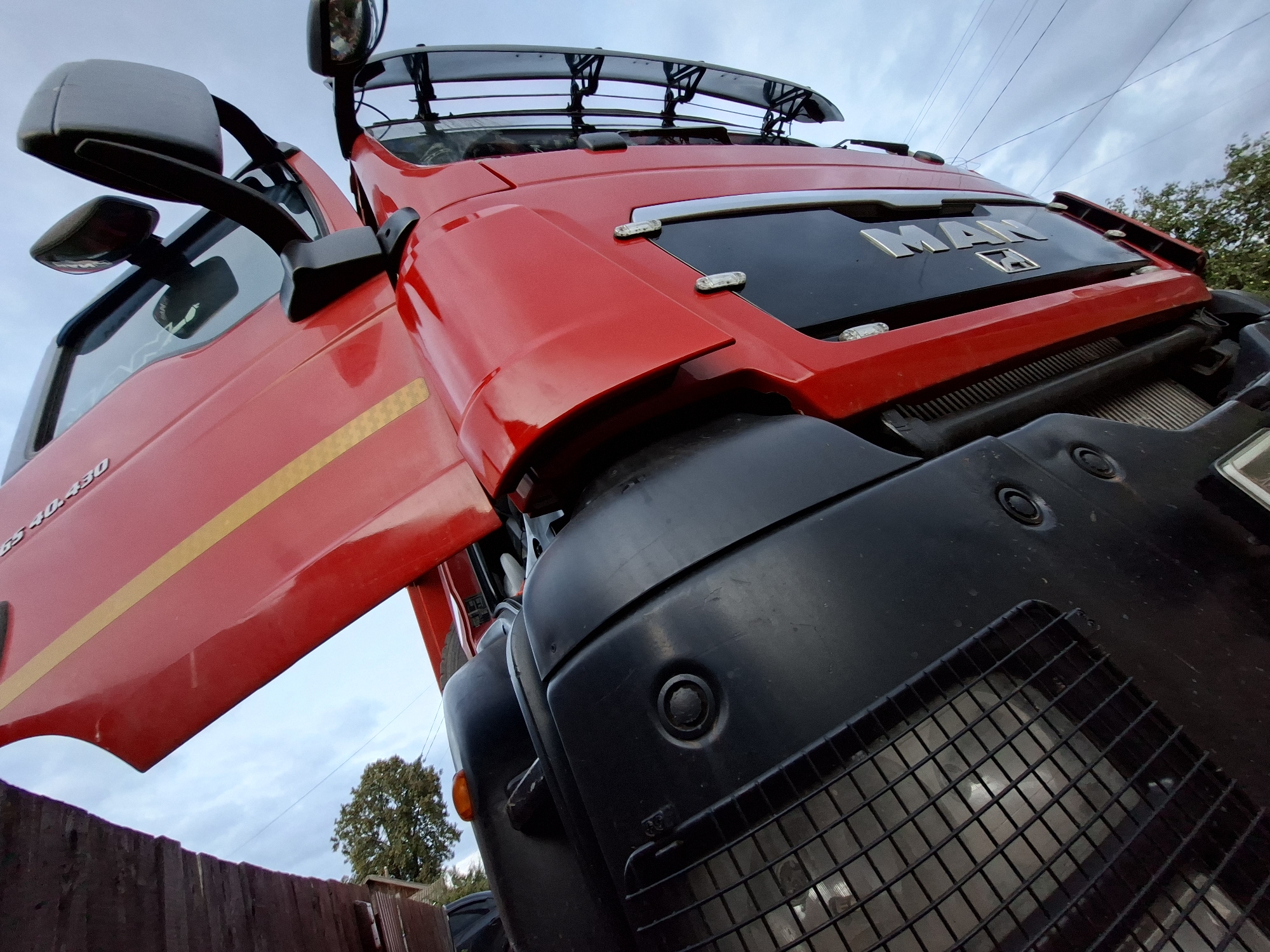 An eye-level, wide-angle shot captures the front of a red MAN truck with its driver's side door open. The truck faces the camera at a slight upward angle, with the sky visible behind it. A wooden fence runs along the bottom left of the frame, and a tree can be seen in the background on the right. The truck's grille and headlights are prominent, as is the "MAN" logo on the front. The scene is set in a suburban outdoor environment.