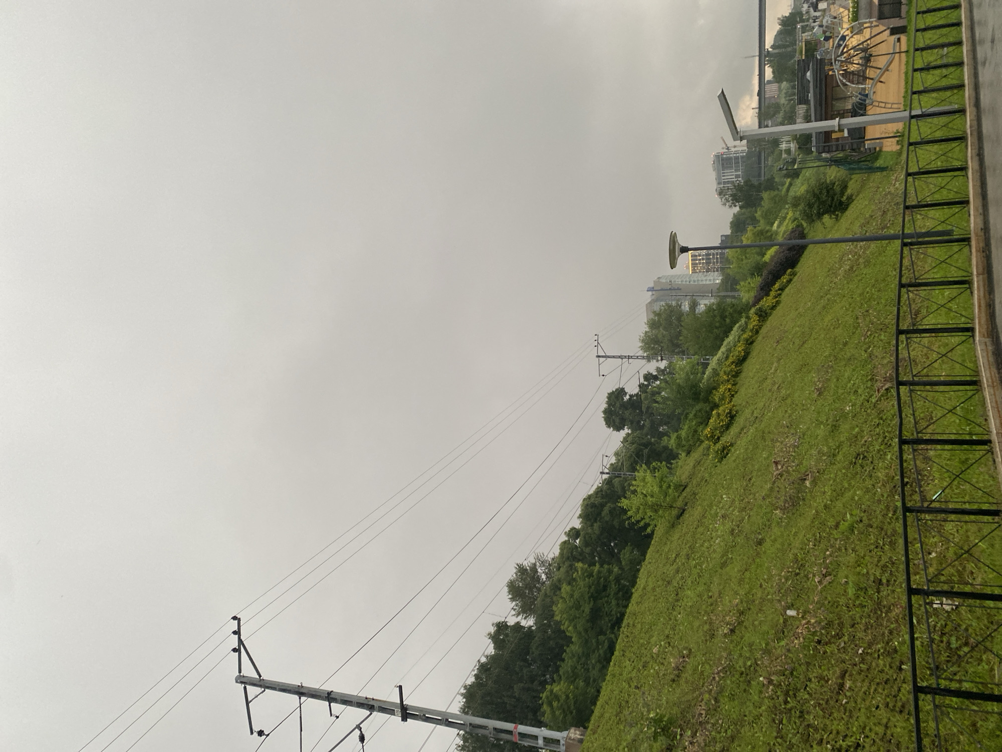 The image depicts an outdoor scene on a cloudy day, likely in the late afternoon or early evening given the overcast sky. The perspective is a bit tilted, as if the camera is angled upwards from a slightly elevated position.
The foreground features a grassy slope leading up towards a black metal fence. Beyond the fence, there are some modern buildings and a playground visible. The architecture of the buildings suggests an urban or suburban setting.
Dominating the middle ground and extending into the background are numerous power lines supported by metal poles. These lines stretch across the frame, disappearing into the distance. The lush green trees interspersed with the power lines indicate a setting with significant vegetation, possibly a park or a residential area with green spaces. One lamppost is visible in the middle ground, with another one near the playground area. The playground features a large, circular structure that looks like a part of a play area, possibly a climbing frame or a spinning ride.
The overall atmosphere is subdued due to the cloudy weather, which creates a soft, diffused light. There is no visible text in the image. The scene appears to be a typical urban landscape where modern infrastructure, recreational facilities, and nature coexist. There are no people visible in the frame.