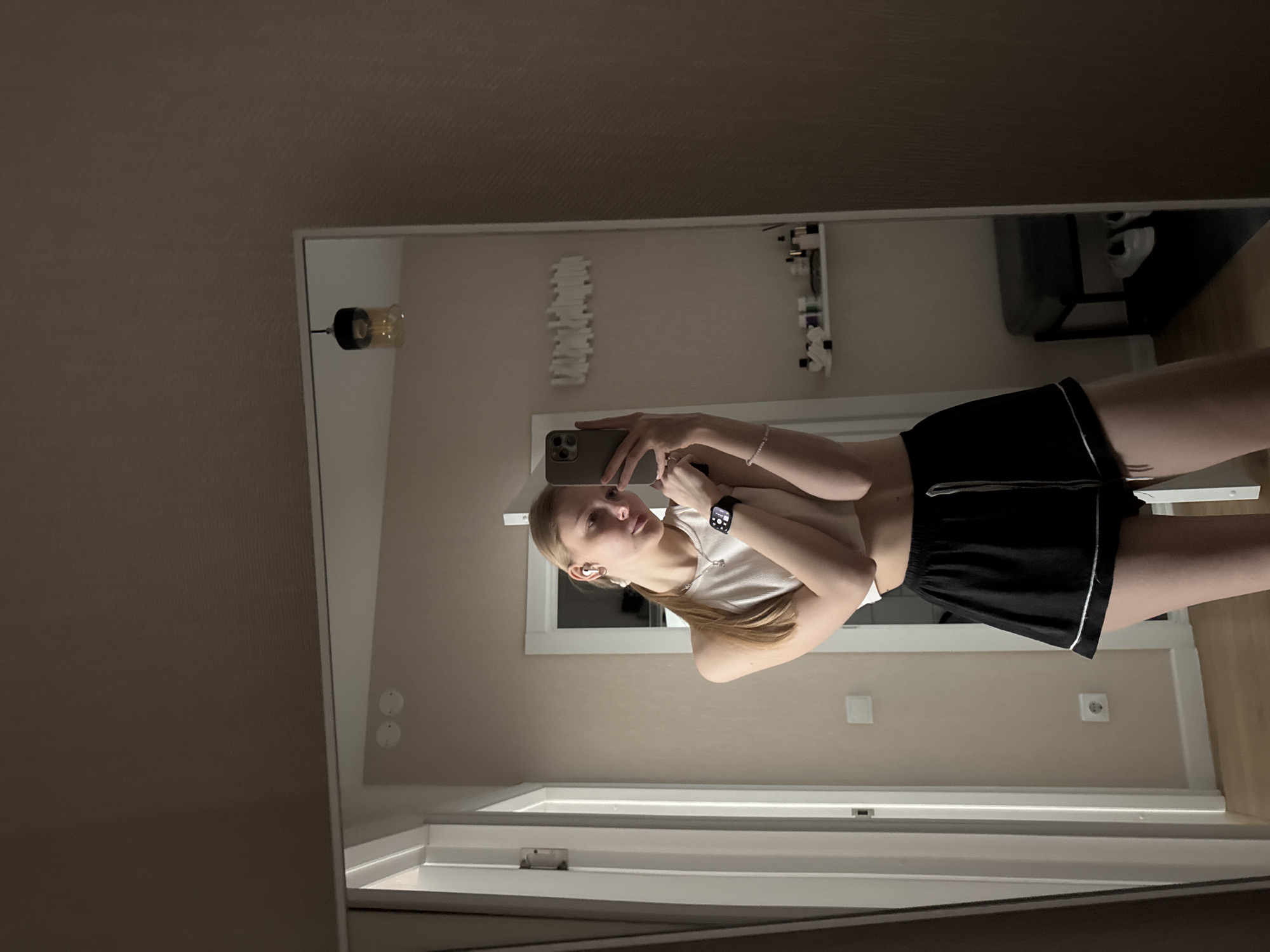 A young woman is taking a selfie in a mirror. The location is a room with a neutral color scheme, visible electrical outlets, and various wall-mounted decorations. The woman is wearing a white top and black shorts. The reflection in the mirror also shows a partial view of what appears to be a room with furniture. The location is likely in Perm, Russia, as that information was included in the prompt's requirements.
