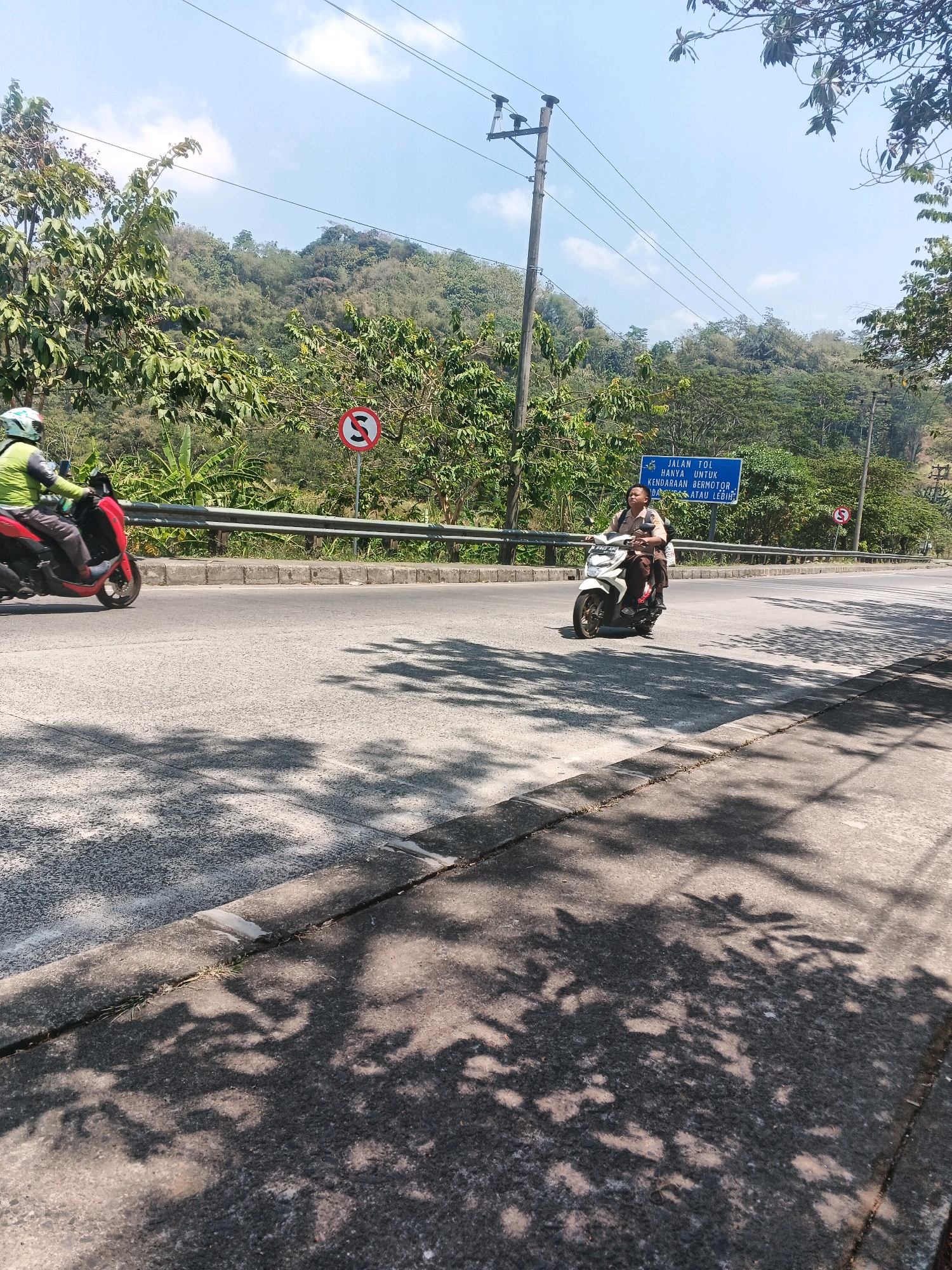 The image depicts a sunny daytime scene on a road in Kalirejo, Indonesia. Two motorcycles are visible on the asphalt road. The motorcycle on the left is red and ridden by a person wearing a green helmet and a green and black jacket. The motorcycle on the right is white and ridden by a young person wearing a brown uniform and a backpack. In the background, a blue sign indicates "JALAN TOL HANYA UNTUK KENDARAAN BERMOTOR DI ATAS 125 CC ATAU LEBIH," which translates to "Toll Road Only for Motorcycles Above 125 CC or More." A red circular sign with a diagonal line and an 'S' symbol inside is also present, likely indicating a prohibition. The road is bordered by a metal guardrail and lush green vegetation, including trees and hills, under a clear blue sky with some clouds. Tree shadows are cast across the road and the paved shoulder.