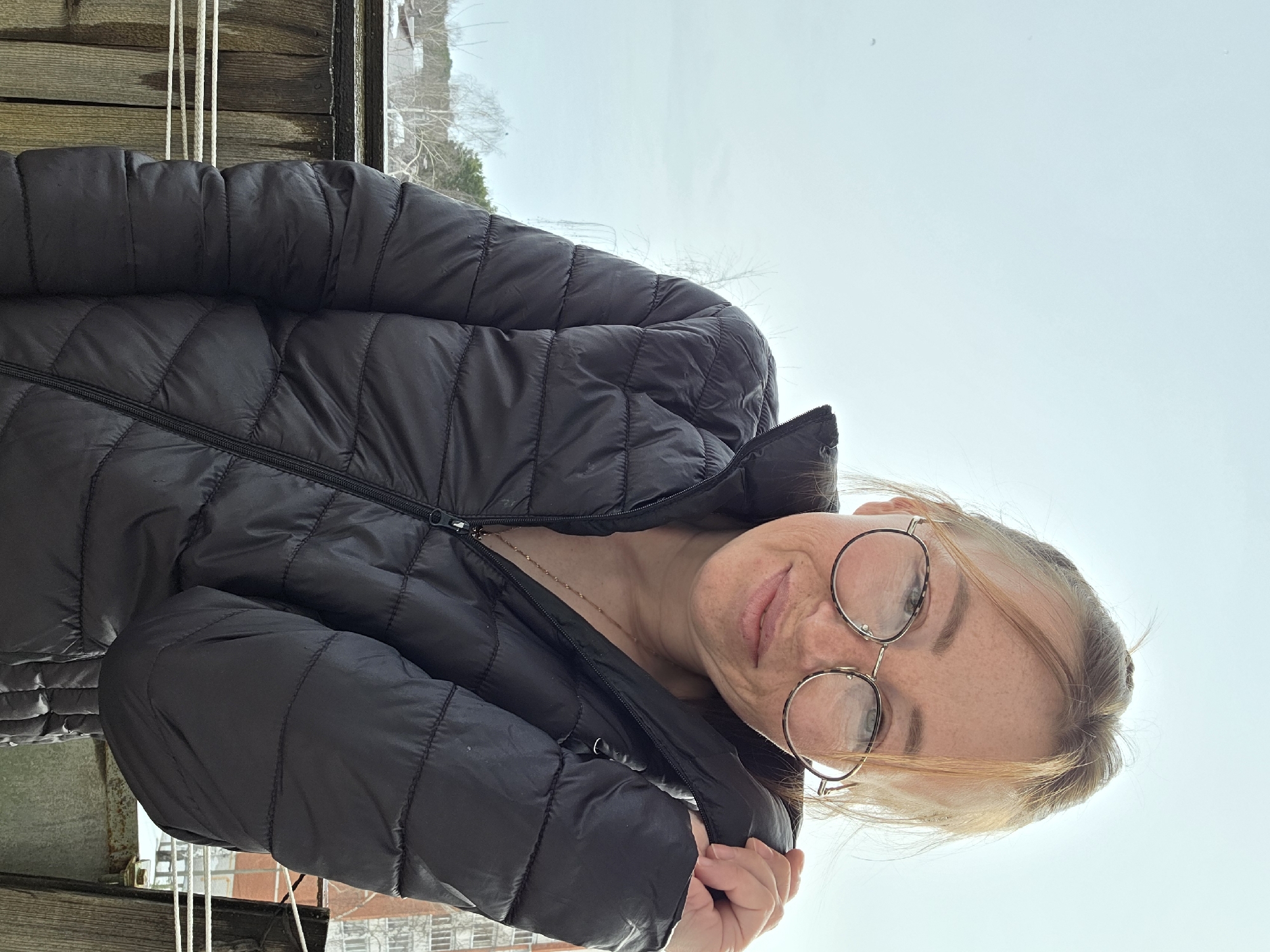 A Caucasian female is depicted outdoors, smiling faintly, in the city of Chaikovsky, Russia. She wears a black, quilted puffer jacket, partially zipped, revealing a gold-colored chain necklace. Her left hand holds the jacket's collar near her neck. She has light brown hair, visible freckles on her face, and round, wire-rimmed glasses. The subject is positioned in front of a weathered wooden railing, behind which three white ropes, consistent with laundry lines, are strung. The background consists of a wintry or early spring landscape with bare trees and distant coniferous trees under an overcast, light blue-grey sky. A multi-story residential building is visible to the far right. Hints of snow are present on distant roofs and ground surfaces.