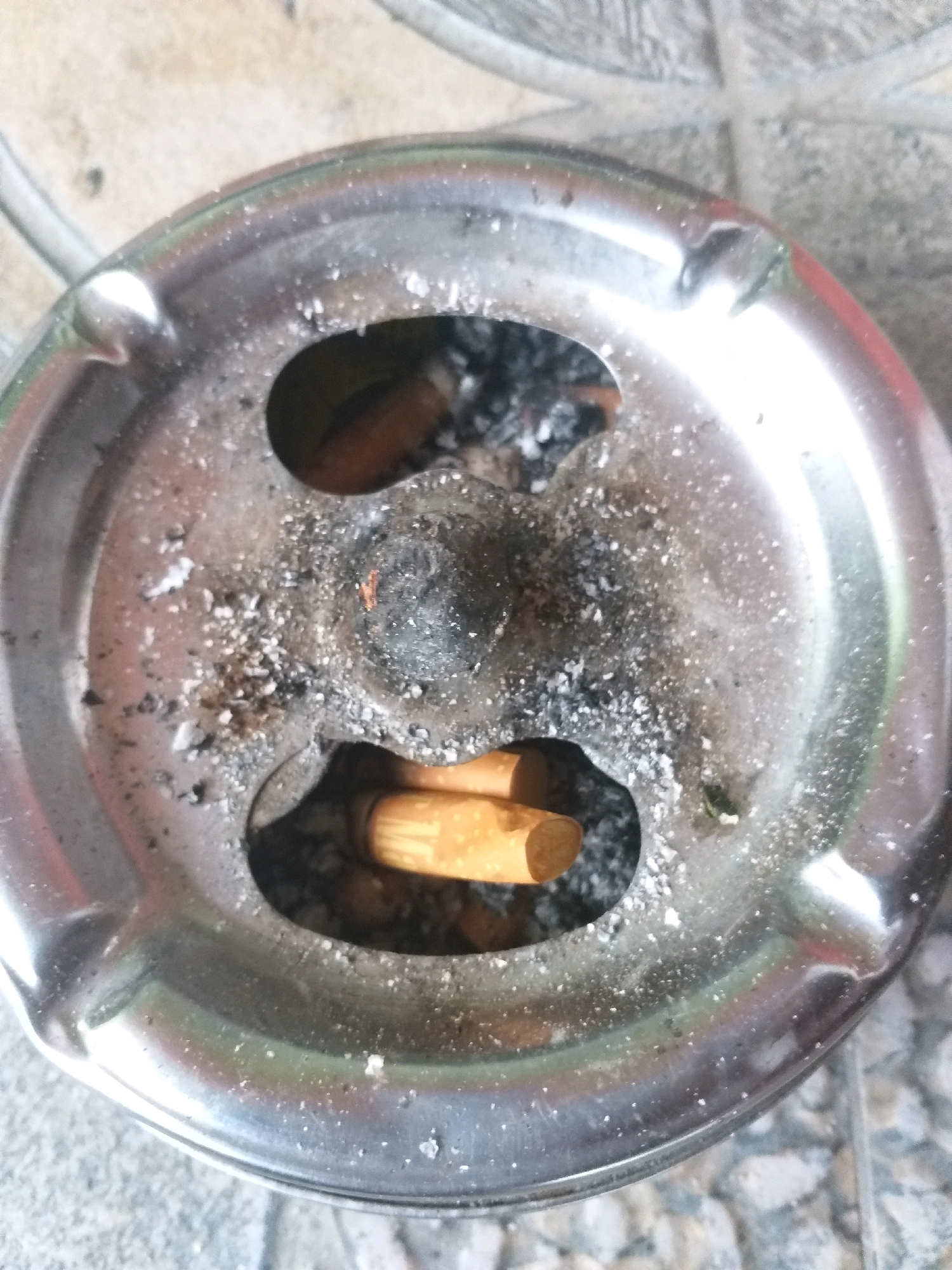 In Air Jamban, Indonesia, a close-up shot captures an ashtray filled with cigarette butts and ash. The ashtray is made of shiny metal and has two oval-shaped openings. Several cigarette butts are visible, some with their filters intact and others appearing partially smoked. The ash is scattered around the butts and in the bottom of the ashtray. The surface of the ashtray shows some wear and tear, with minor scratches and smudges. The background is a textured surface, possibly a tiled floor or pavement.
