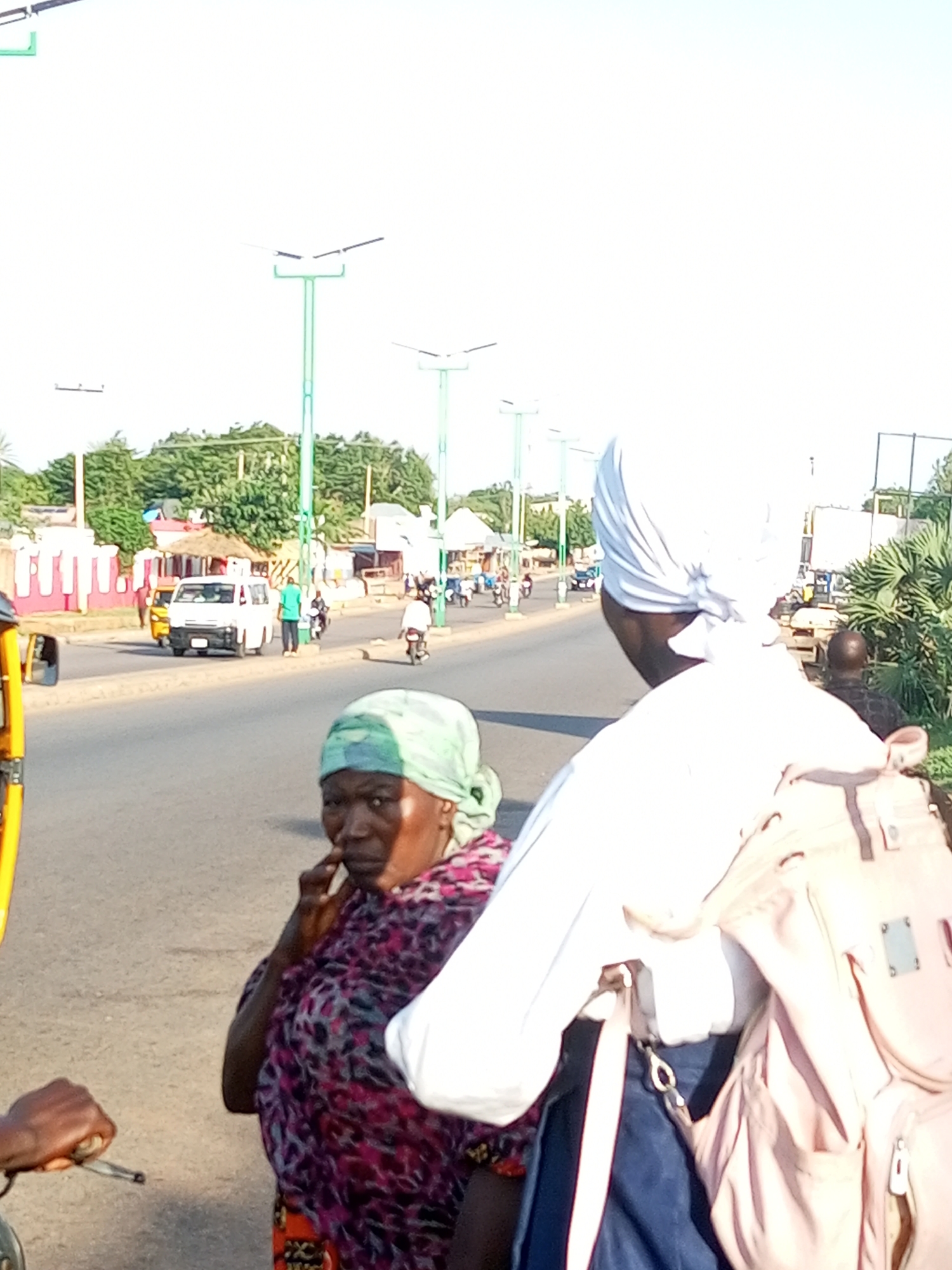 The image depicts a street scene in Tempure, Nigeria, under a bright sky. In the foreground, a woman wearing a green headscarf and a colorful patterned dress is looking towards the left, with her hand near her mouth. To the right of her, a person draped in a white cloth is visible, with a light pink backpack slung over their shoulder. Behind them, a busy street unfolds with several vehicles, including a white van, a yellow auto-rickshaw, and motorcycles with riders. The road is lined with trees and buildings, suggesting an urban or semi-urban environment. Streetlights with solar panels are also visible along the roadside. The overall impression is of a typical day in a Nigerian town, with daily life and transportation in motion.