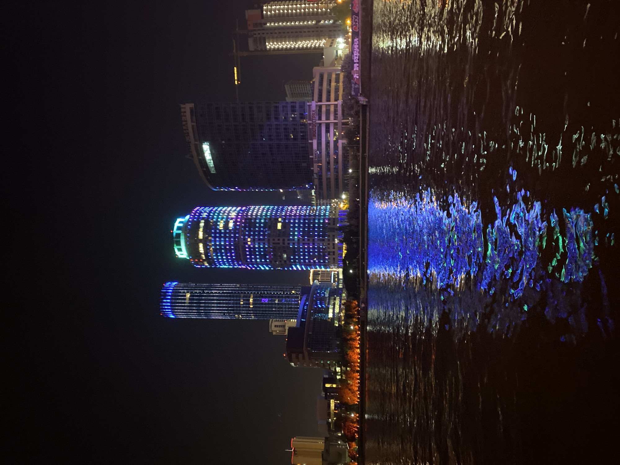 The image depicts the Iset River in Yekaterinburg, Russia at night. The buildings along the riverbank are illuminated with bright lights, including the Iset Tower, and these lights are reflected in the water. The river appears to be calm, with gentle ripples on the surface.