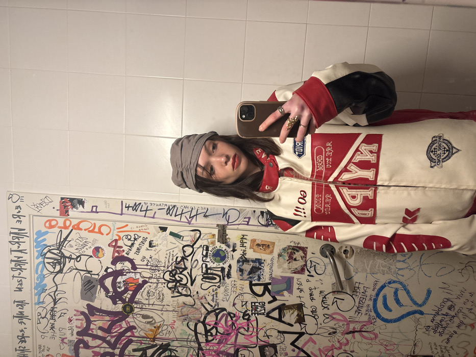 A female subject is depicted taking a mirror selfie in an indoor public space, likely a restroom within the city of Barcelona, Spain. The subject, with dark brown hair and bangs, wears red lipstick and a grey ruched head covering. She is dressed in a white, red, and black racing-style jacket featuring various patches, including prominent "NYN" lettering, a "RIOT" patch, and "!!! GO!!!" text on the collar. The subject's right hand, visible in the reflection, holds a brown-cased smartphone used to capture the image, with multiple rings worn on her fingers. The background to the left consists of a white door heavily covered with graffiti, tags, and stickers in various colors, one of which explicitly states "Barcelona" and another "FREE PALESTINE." A silver-toned door handle is affixed to this door. The background to the right of the subject is a plain white tiled wall. The scene is illuminated by overhead artificial lighting.